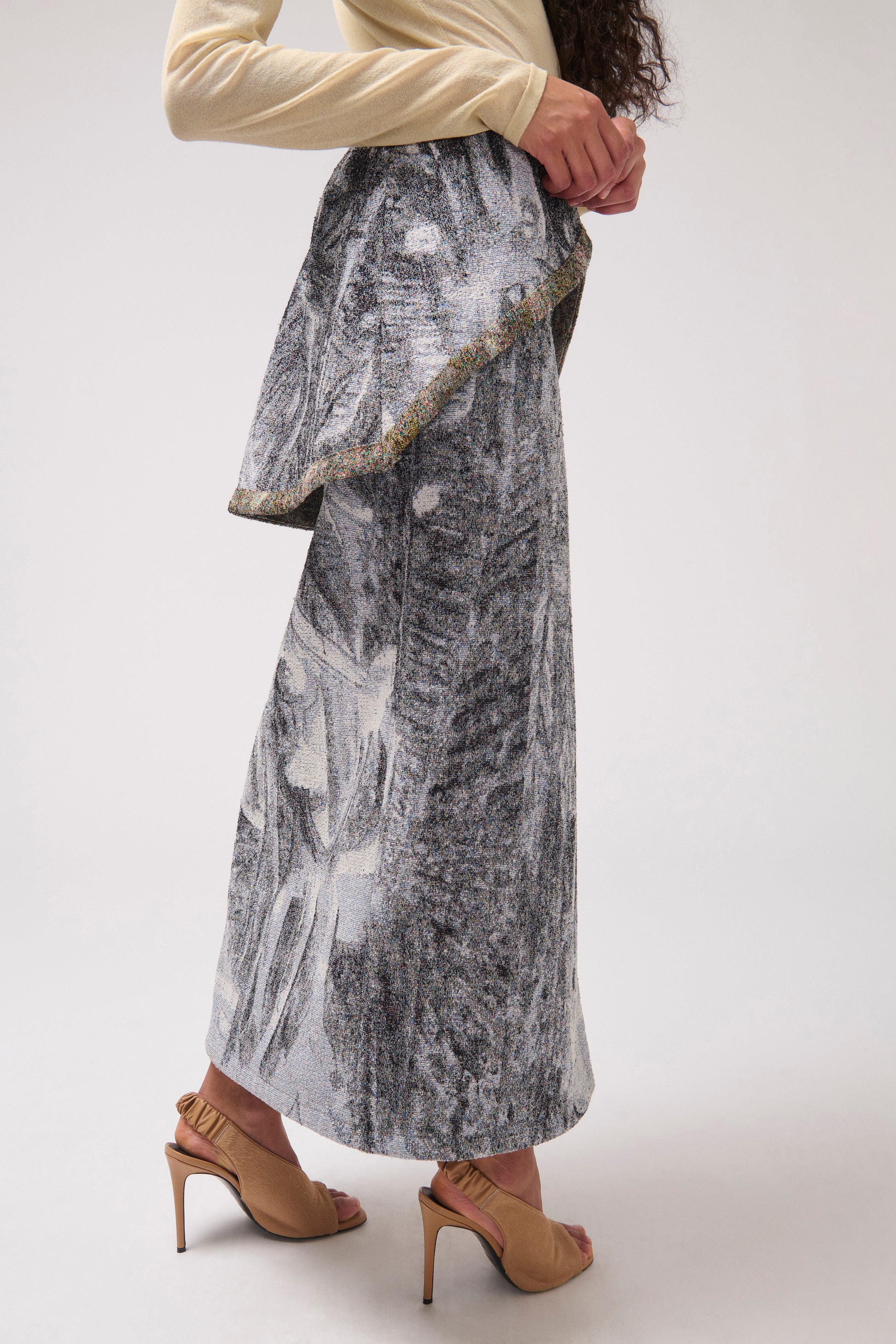 Back view of a model standing against a light gray studio background wearing a  long draped skirt with a multi-color print. and beige heeled sandals.
