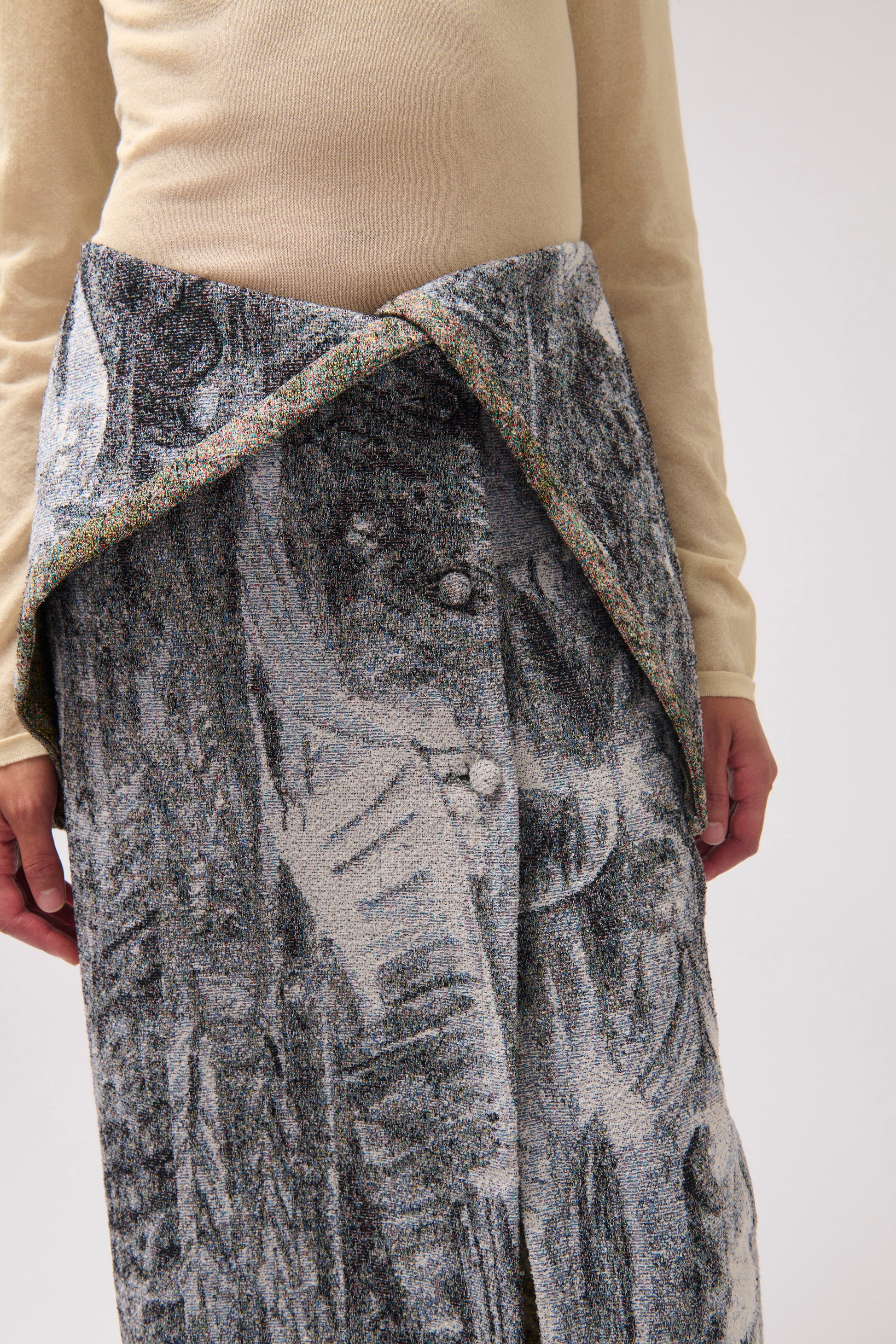A close-up of a person in front of a light gray background wearing a long draped skirt with a multi-color print. 