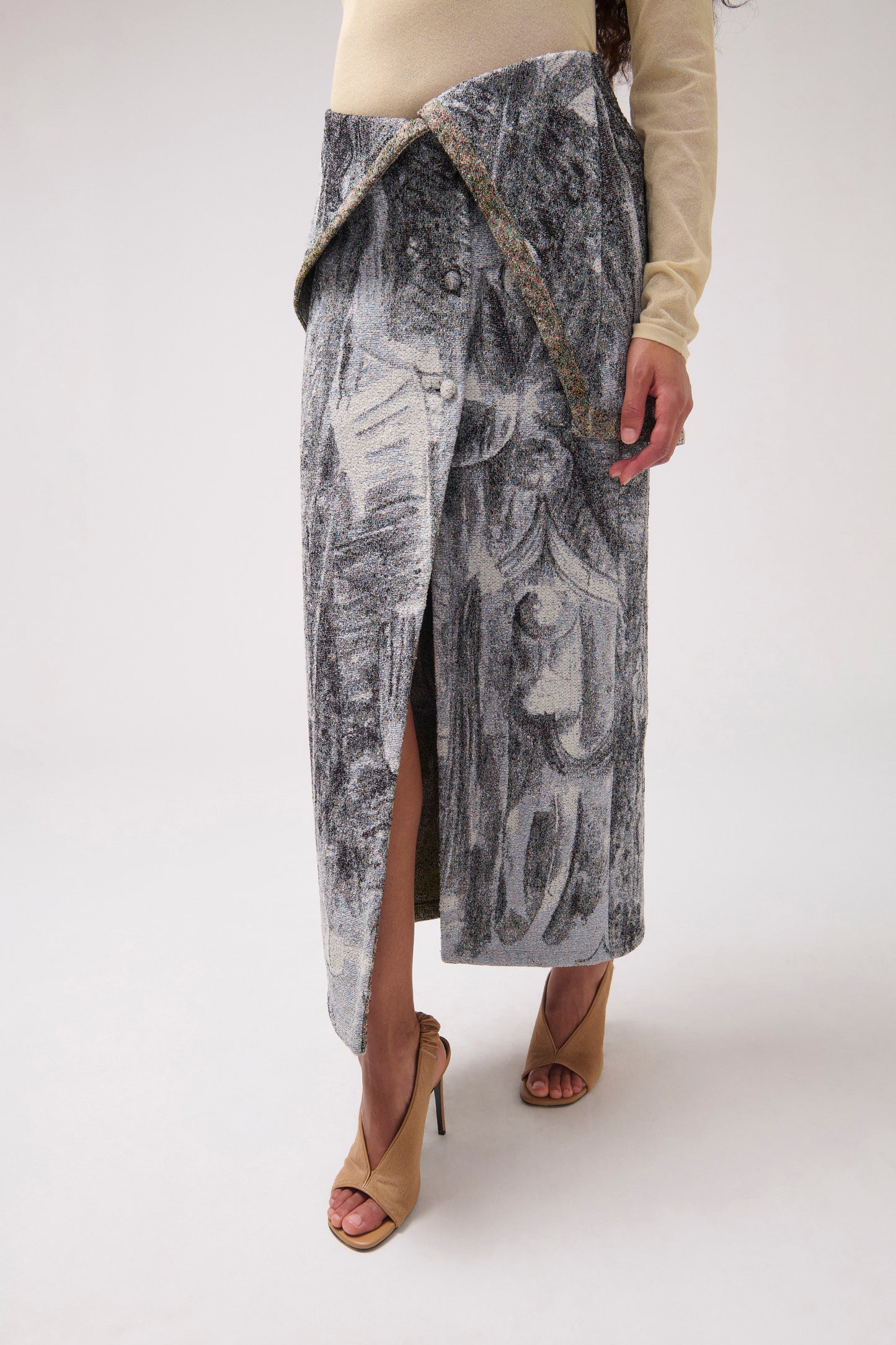 A close-up of model stands against a light gray studio background wearing a  long draped skirt with a multi-color print. 