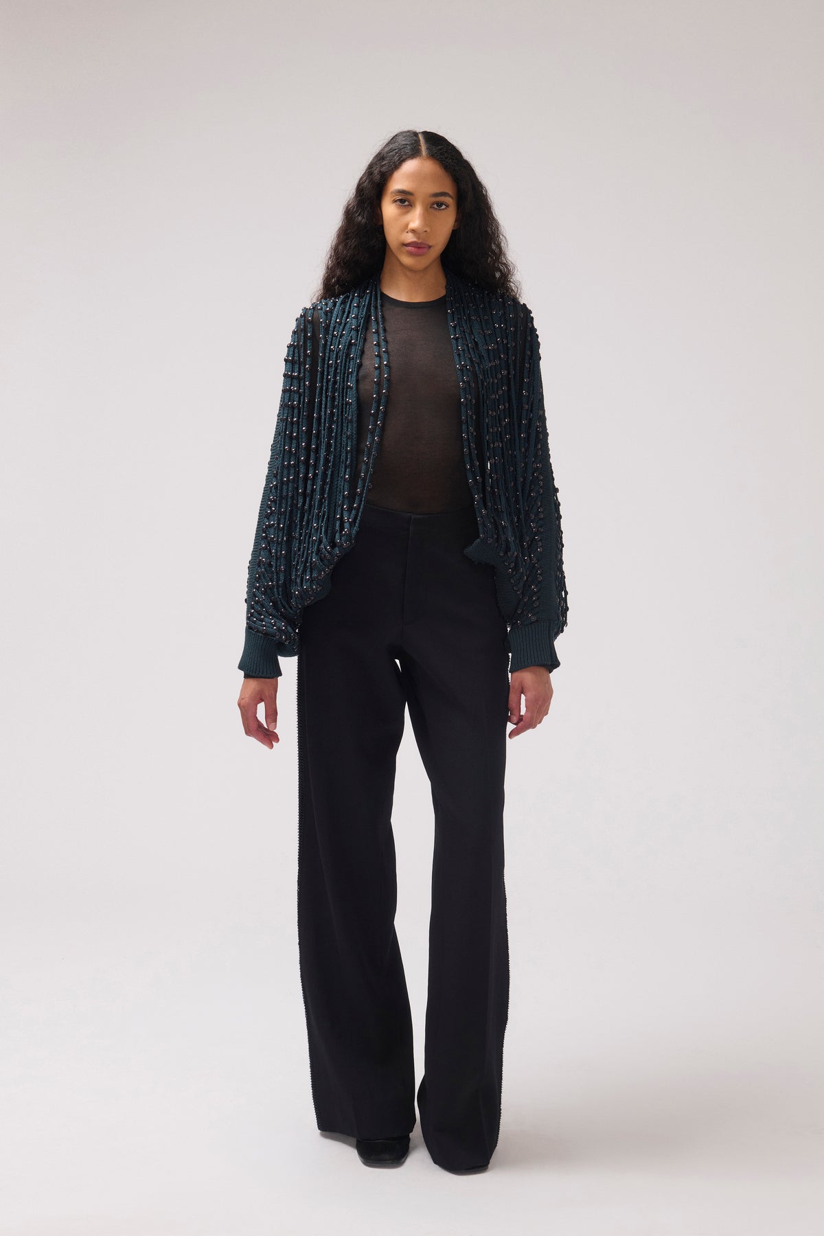 A model stands against a light gray studio background wearing a dark teal beaded open-front jacket over a black top and black wide-leg trousers.