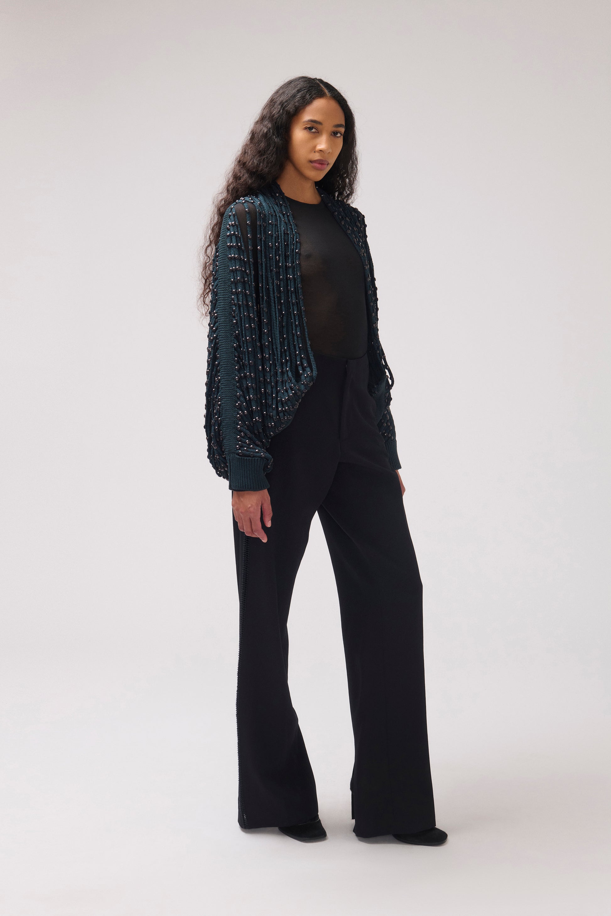 A model stands in a studio wearing a dark teal textured, beaded cardigan over a black top and black wide-leg trousers, photographed against a light gray background.