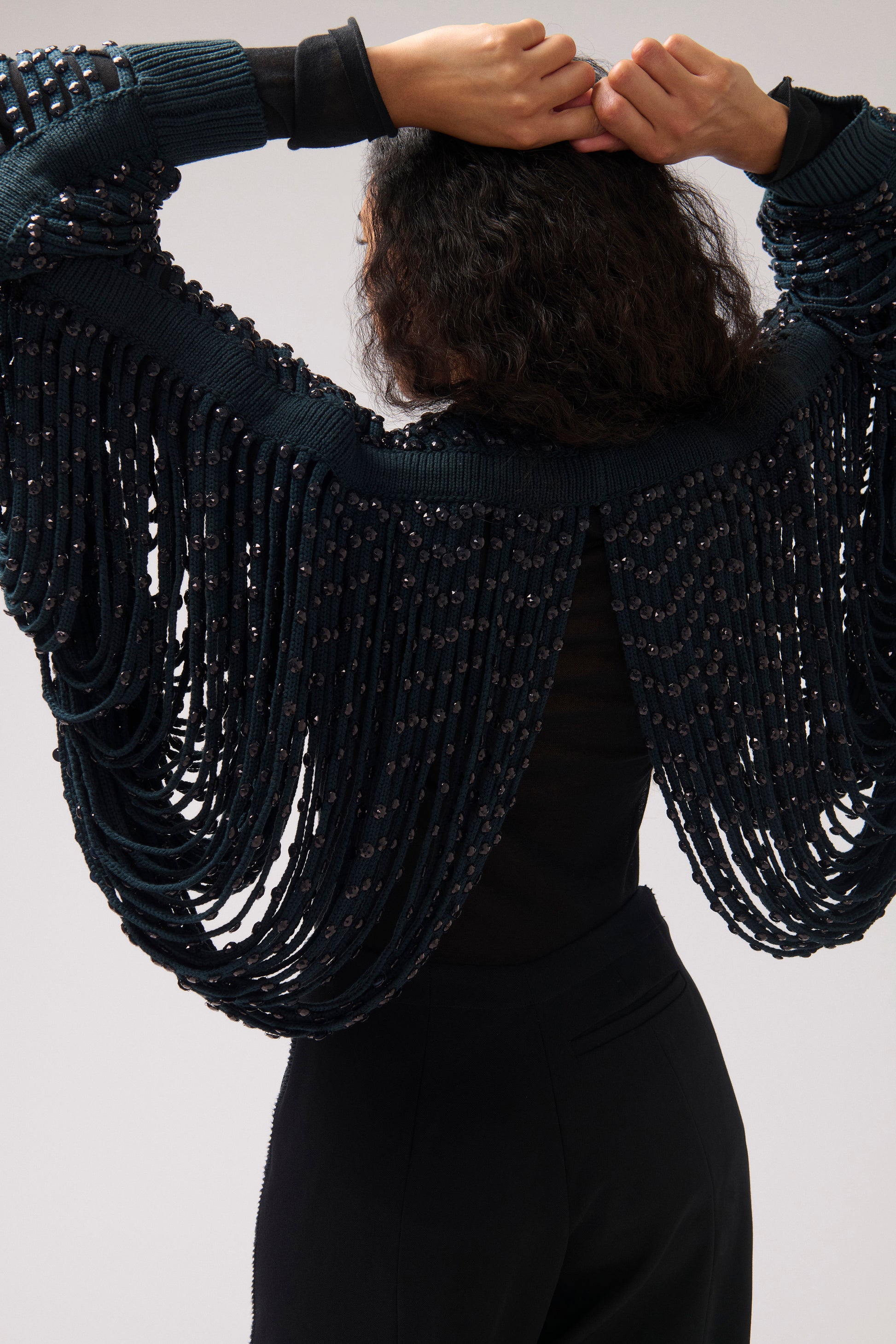 A model from behind wearing a dark navy blue knit shrug or cape with cascading fringe and bead details, paired with a black top and black pants, against a light gray background.