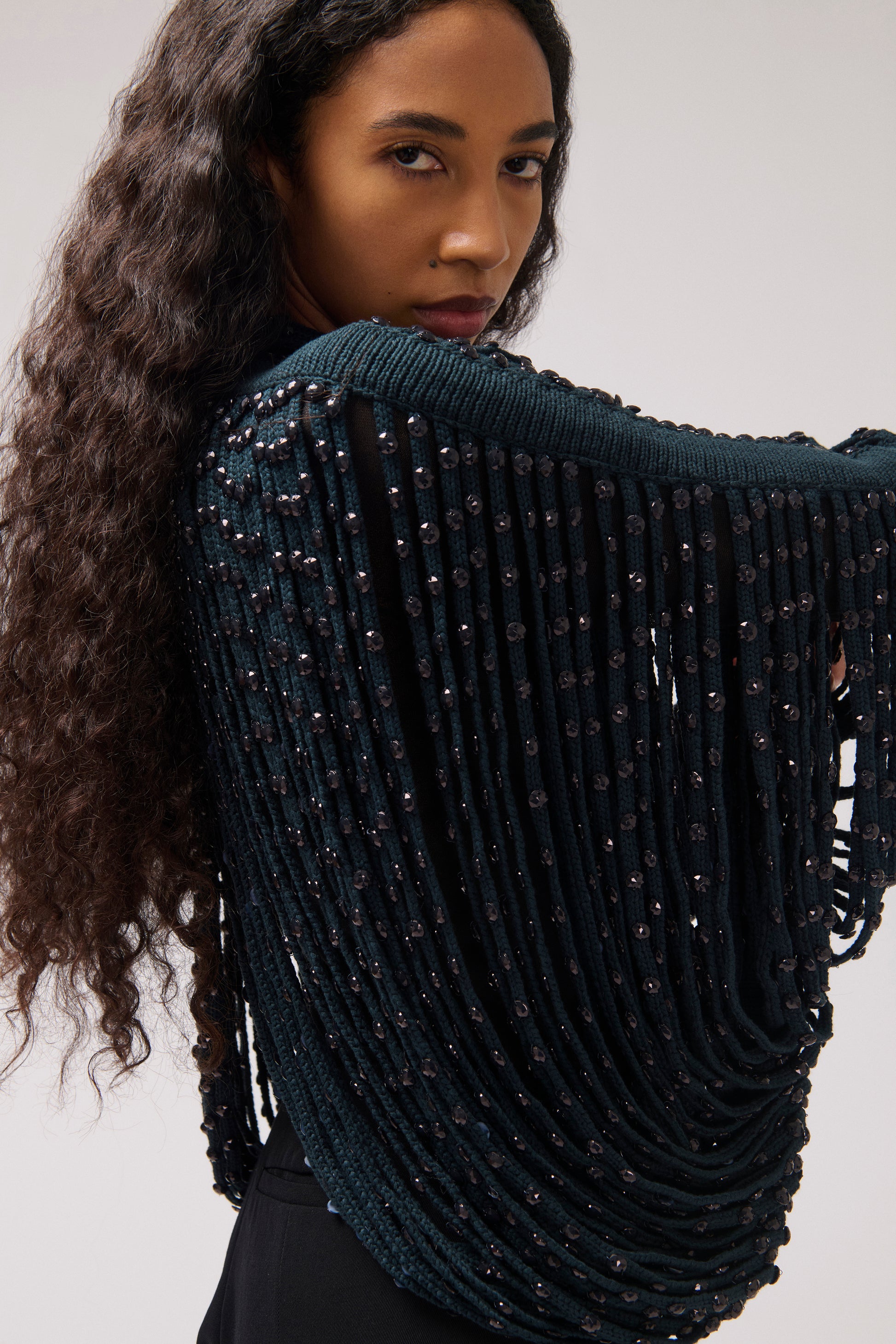 A woman wearing a dark teal fringe knit top with beaded details, shown from the back against a light gray studio background.