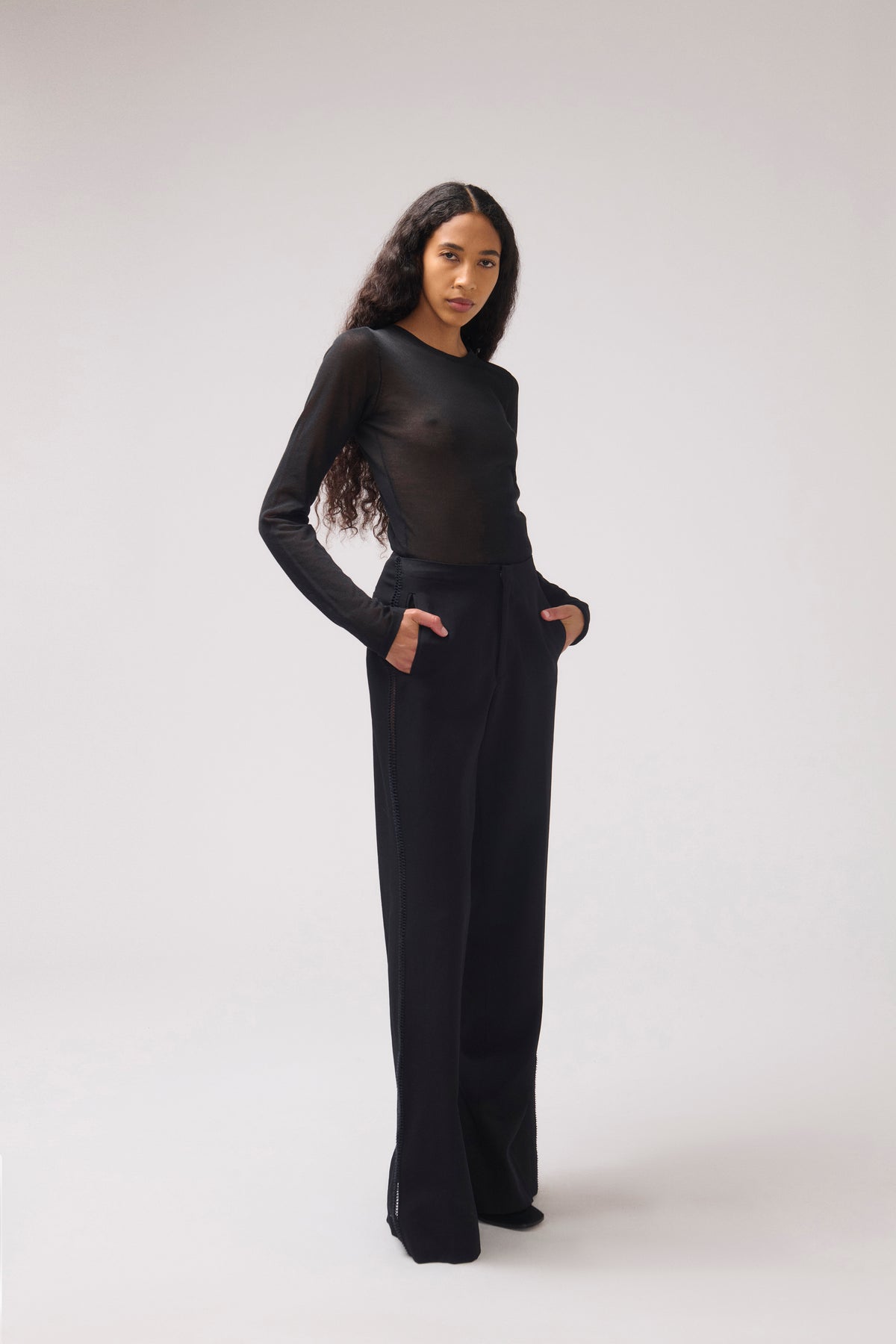 A model modeling a black sheer long-sleeve top paired with black trousers, standing against a light gray studio background.