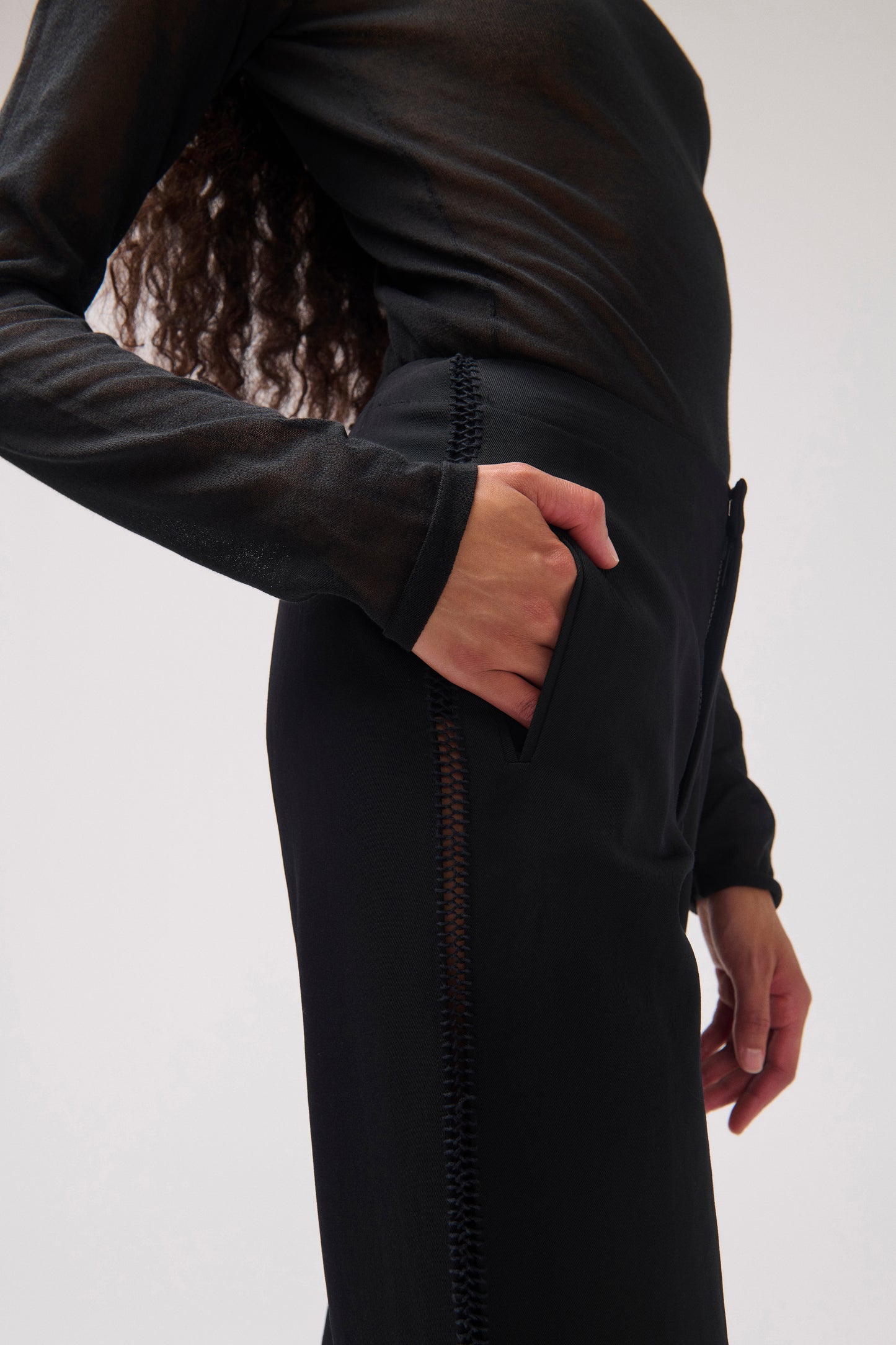 A close-up side view of a person wearing a black sheer long-sleeve top and a black wool pant with a vertical stitched detail, against a plain light gray background.