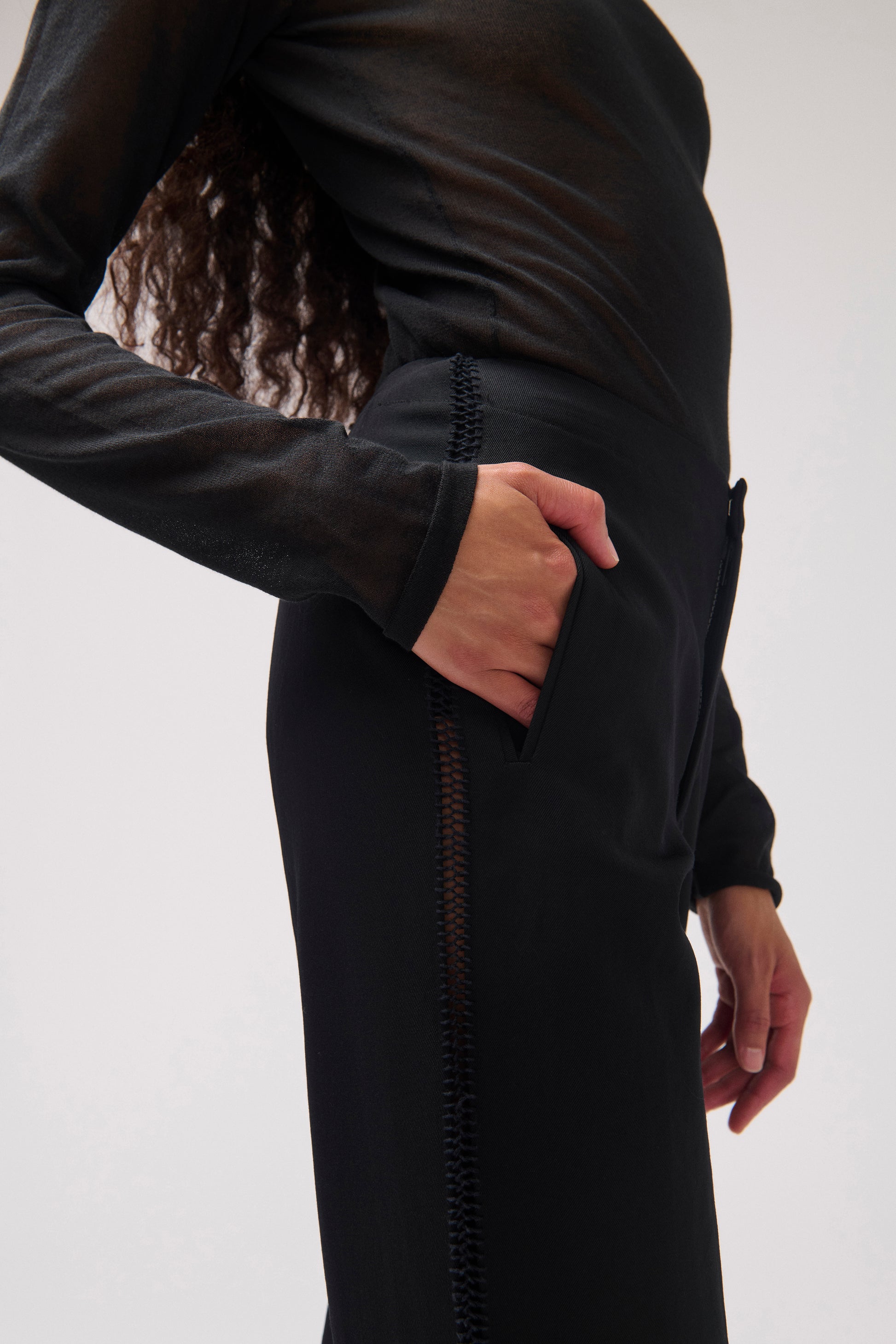 A close-up side view of a person wearing a black sheer long-sleeve top and a black wool pant with a vertical stitched detail, against a plain light gray background.