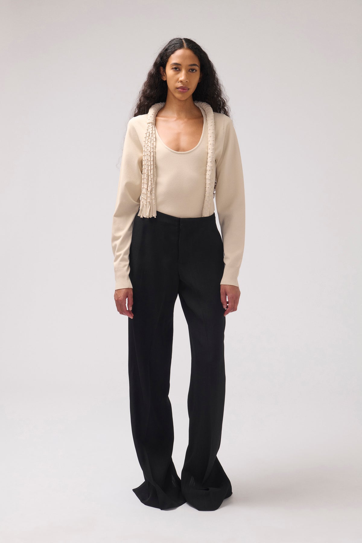 A model stands against a light gray studio background wearing a cream long-sleeve top with decorative fringe trim and black wide-leg trousers.