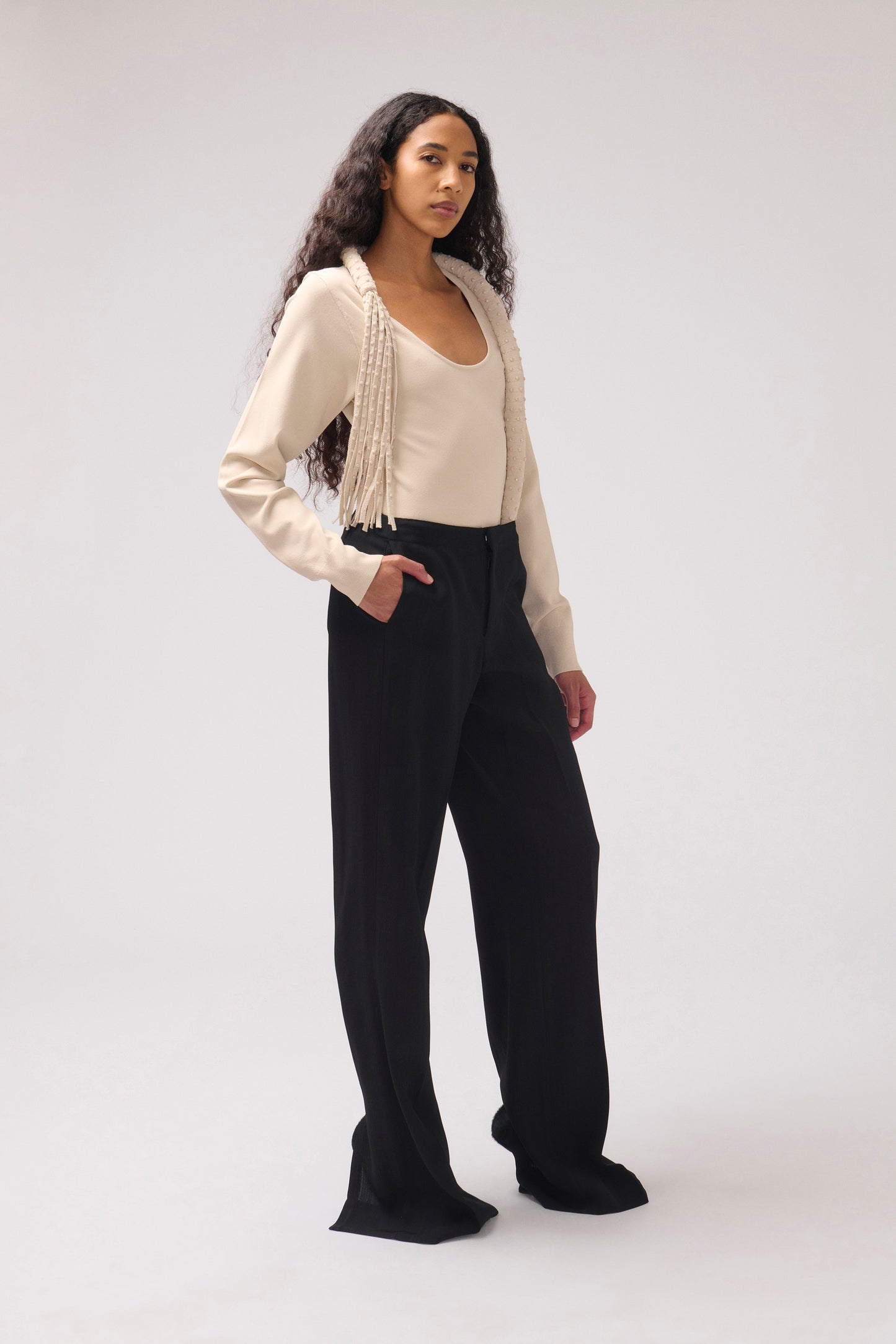 A model stands against a light gray studio background wearing a cream long-sleeve top with fringe detail and black wide-leg pants.