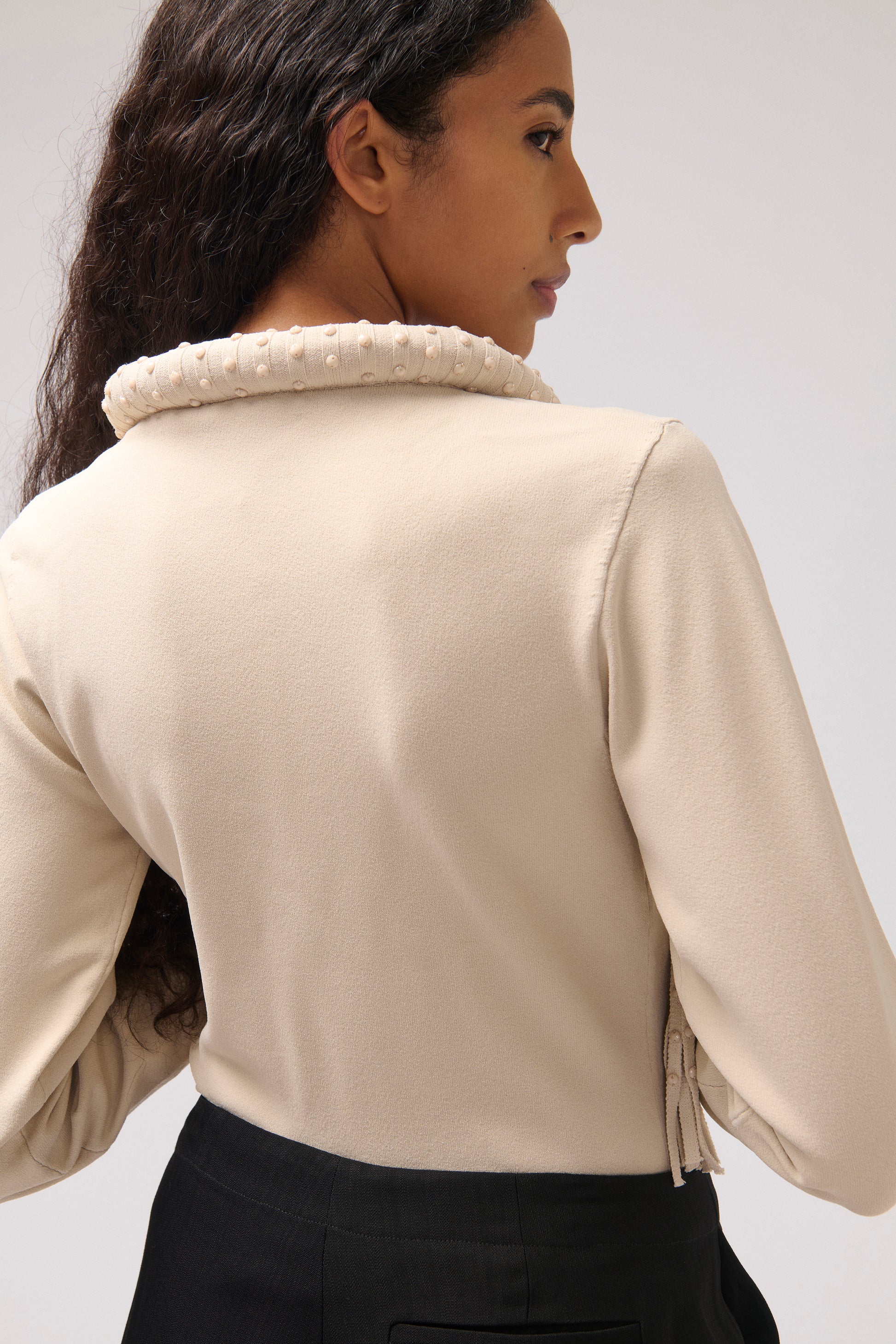 A woman shown from the back wearing a beige long-sleeve blouse with a textured, pearl-like collar and black bottoms, photographed against a light gray studio background.