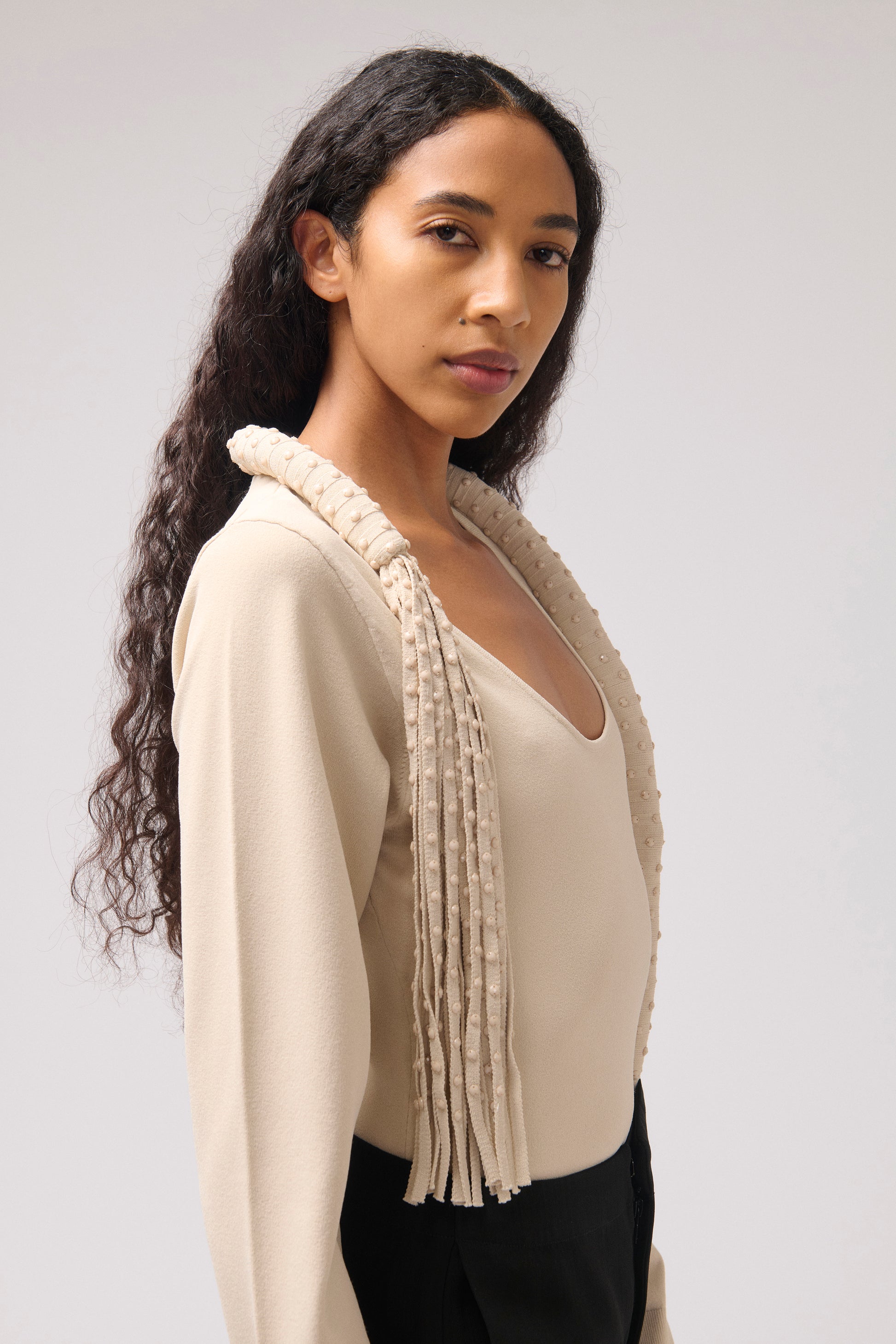 A woman modeling a beige knit top with a wide, fringed textured collar, paired with black bottoms, against a neutral light gray background.
