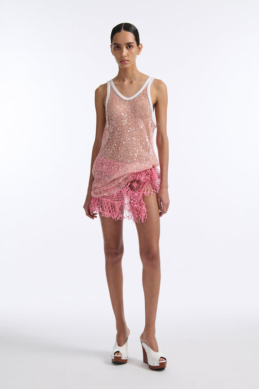 Female model with slicked back black hair in a bun wears pink iridescent see-through sequin tank top with white piping. Model also wears see-through bright pink crochet skirt and white wedges.