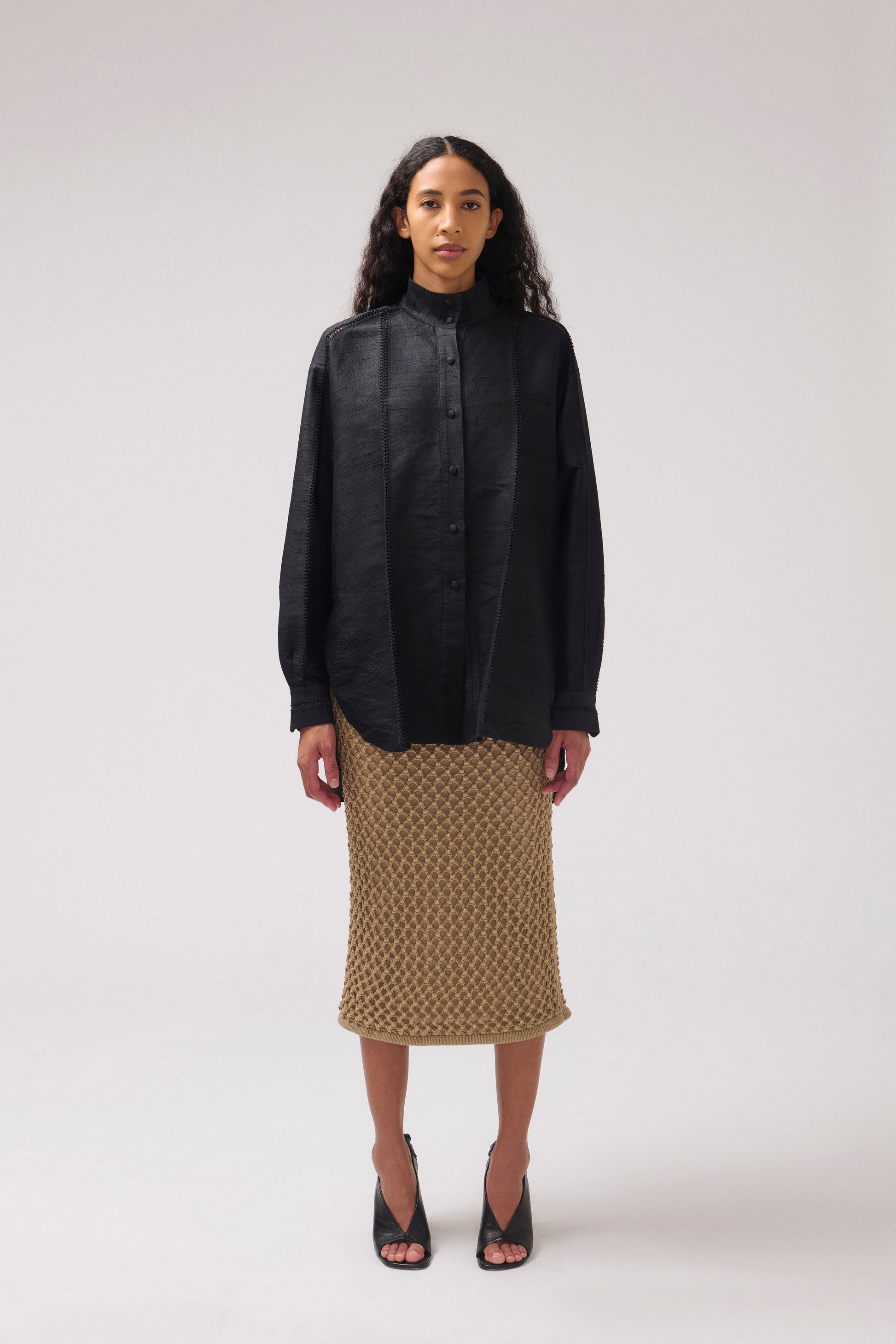 A model facing the camera is standing against a light gray studio background wearing a black oversized long-sleeve silk shirt and a textured knit midi skirt with black high-heeled sandals.