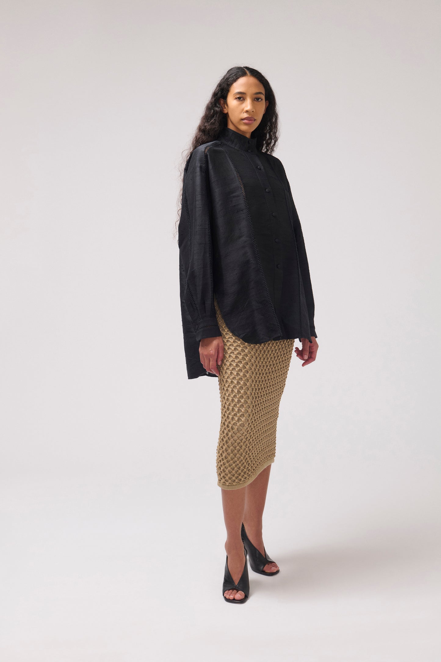 A model stands against a light gray studio background wearing a black oversized long-sleeve silk shirt and a textured knit midi skirt with black high-heeled sandals.
