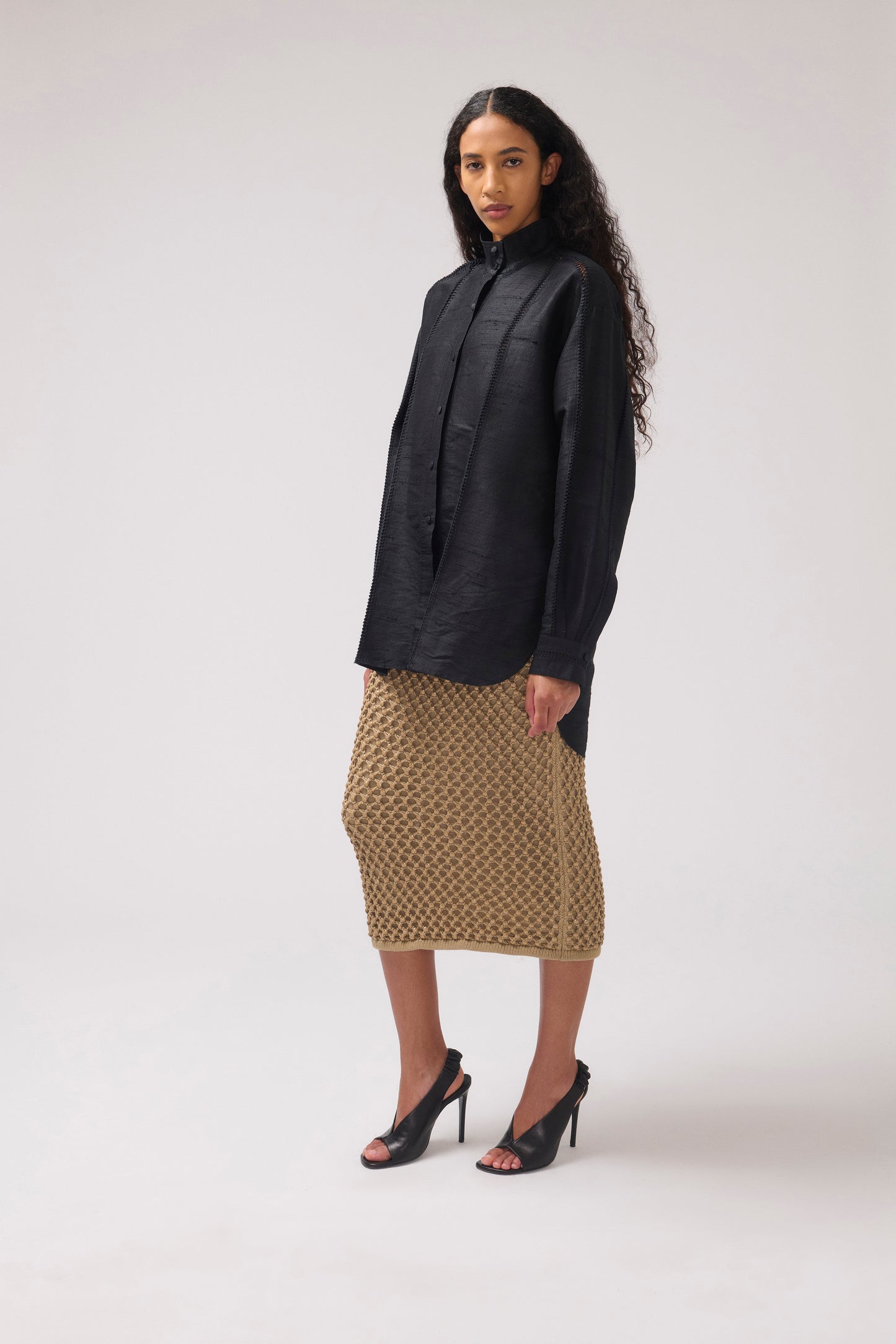 A model stands in a studio wearing a black long-sleeve button-up shirt and a textured midi skirt with black high heels, photographed against a light gray background.