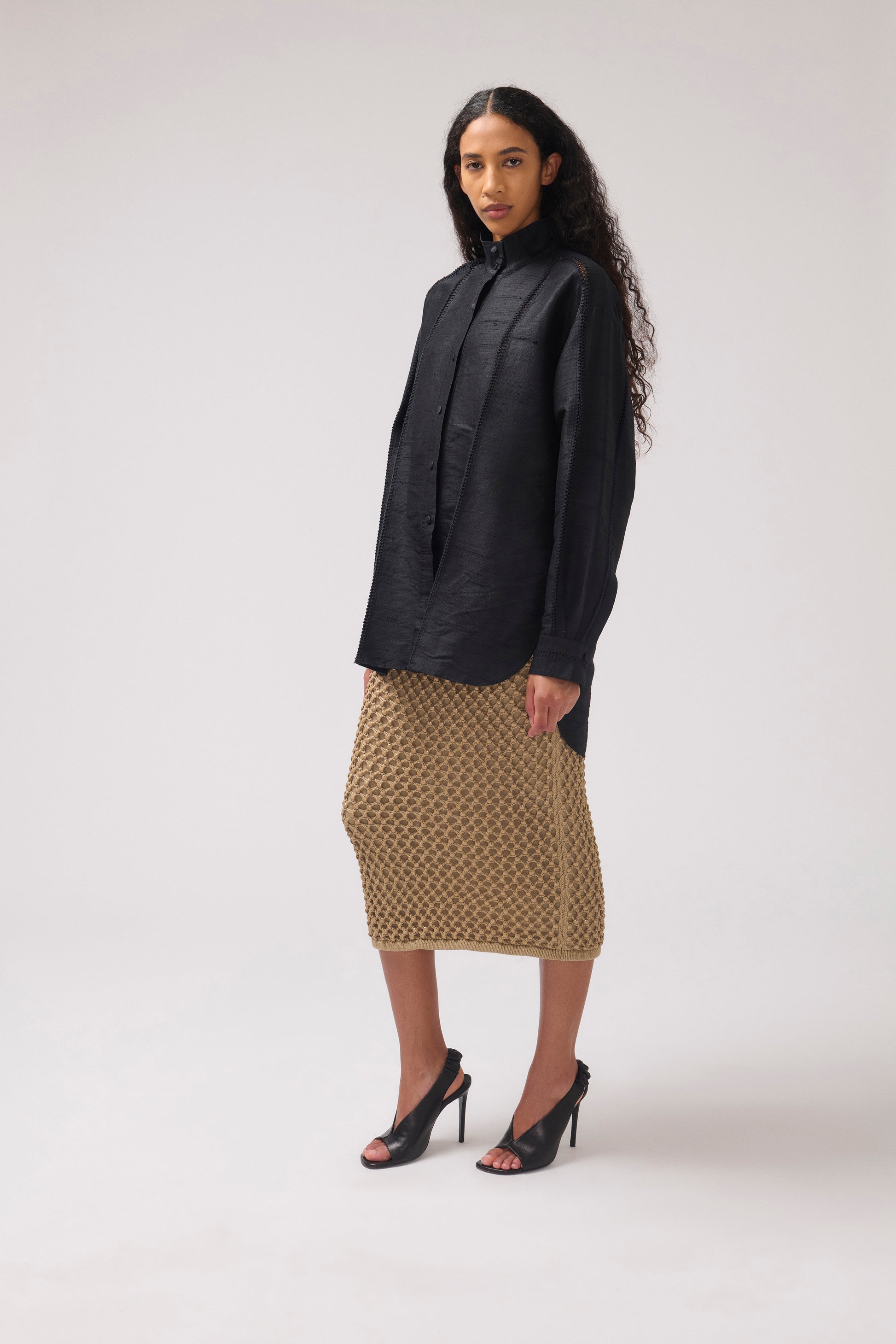 A model stands in a studio wearing a black long-sleeve button-up shirt and a textured midi skirt with black high heels, photographed against a light gray background.