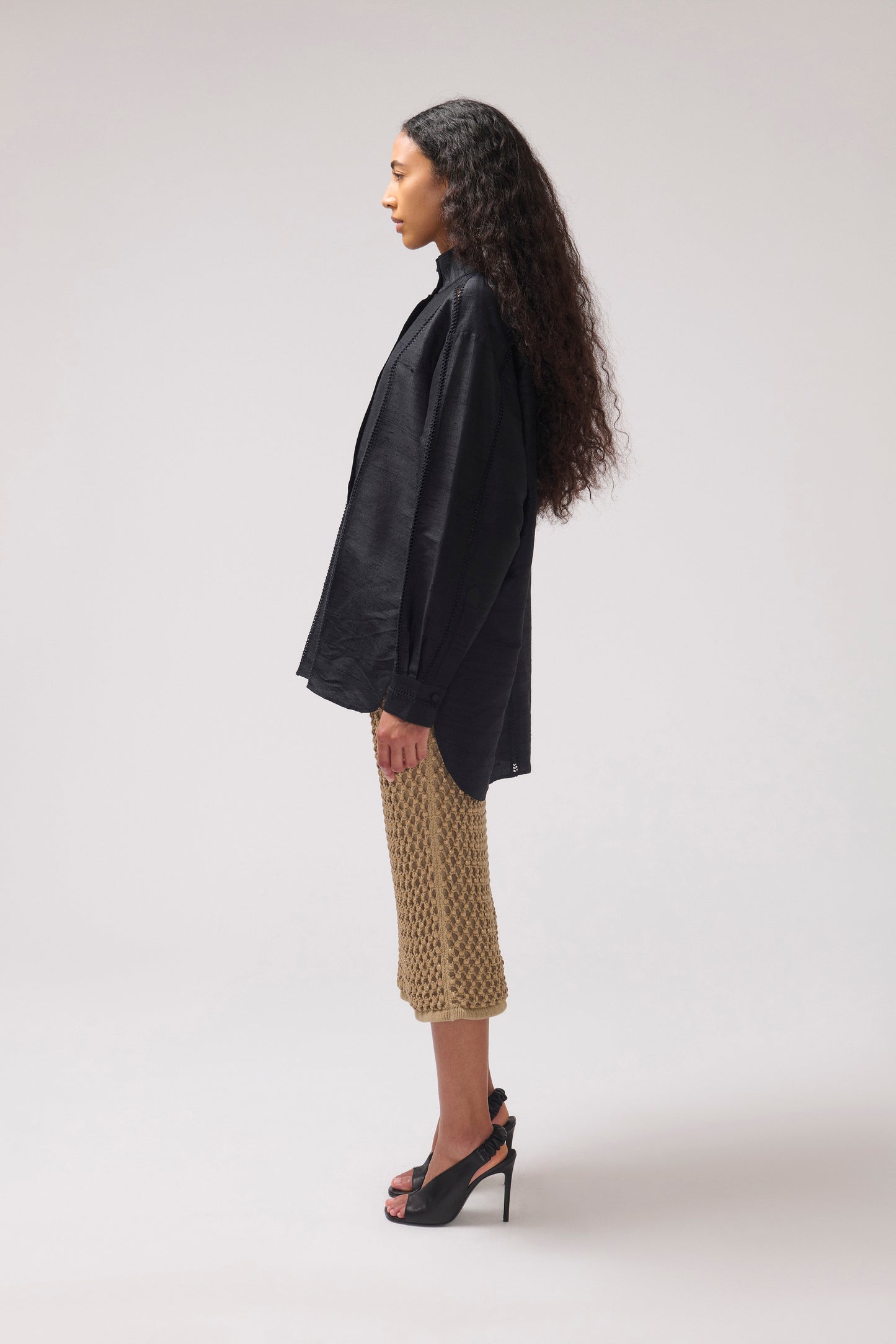 A woman stands in profile wearing a loose black long-sleevesilk shirt, a midi skirt, and black high-heeled sandals against a plain light gray studio background.