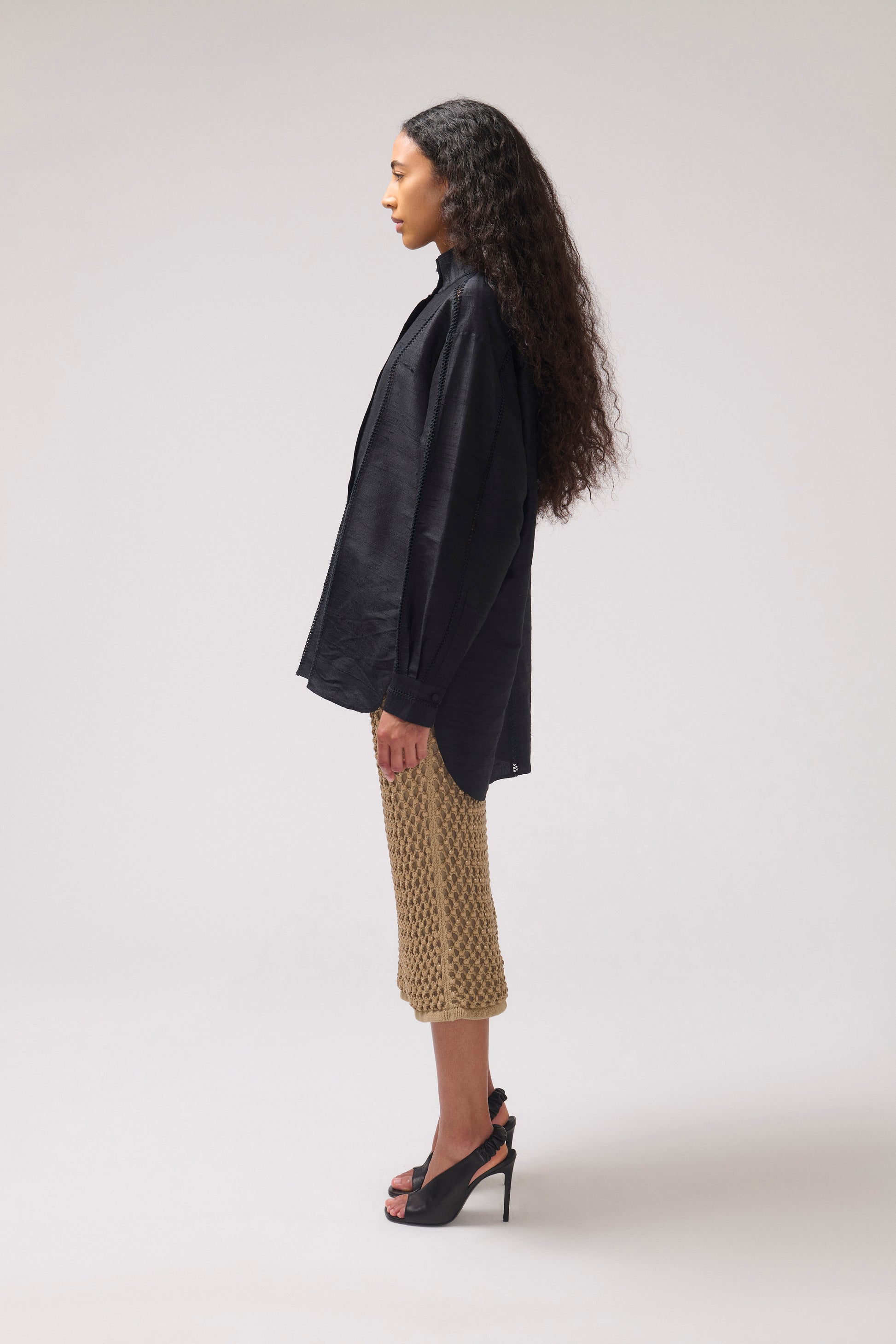 A woman stands in profile wearing a loose black long-sleevesilk shirt, a midi skirt, and black high-heeled sandals against a plain light gray studio background.