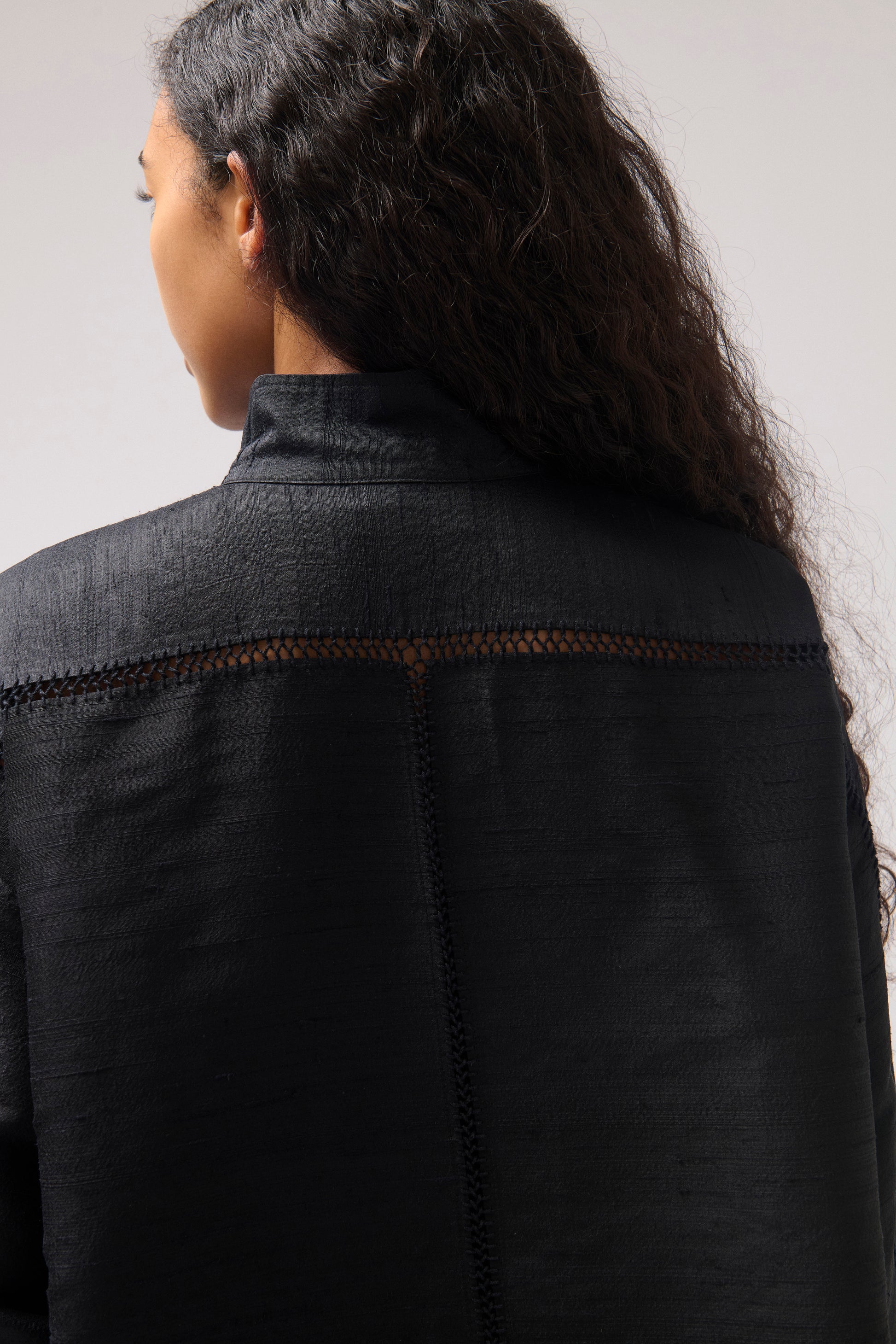 Close-up back view of a person wearing a black texturedsilk shirt with a high collar and decorative hand-made stitching across the shoulders, against a plain light gray background.