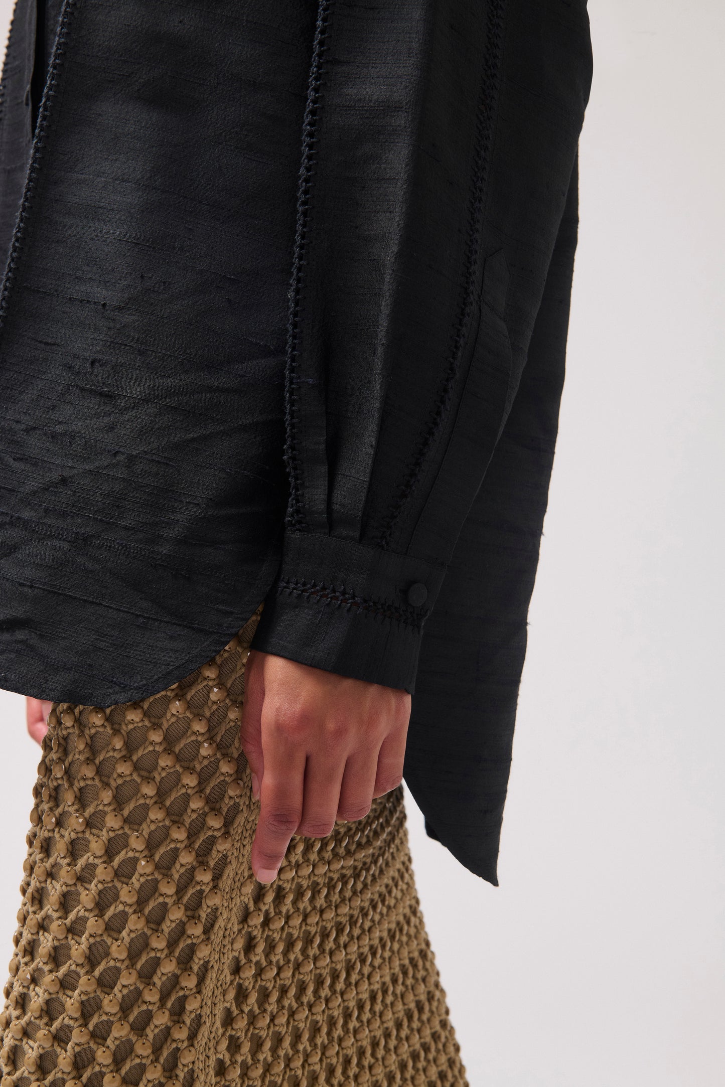 Close-up of a person wearing a black textured long-sleeve blouse with a button cuff and a skirt against a light gray background.