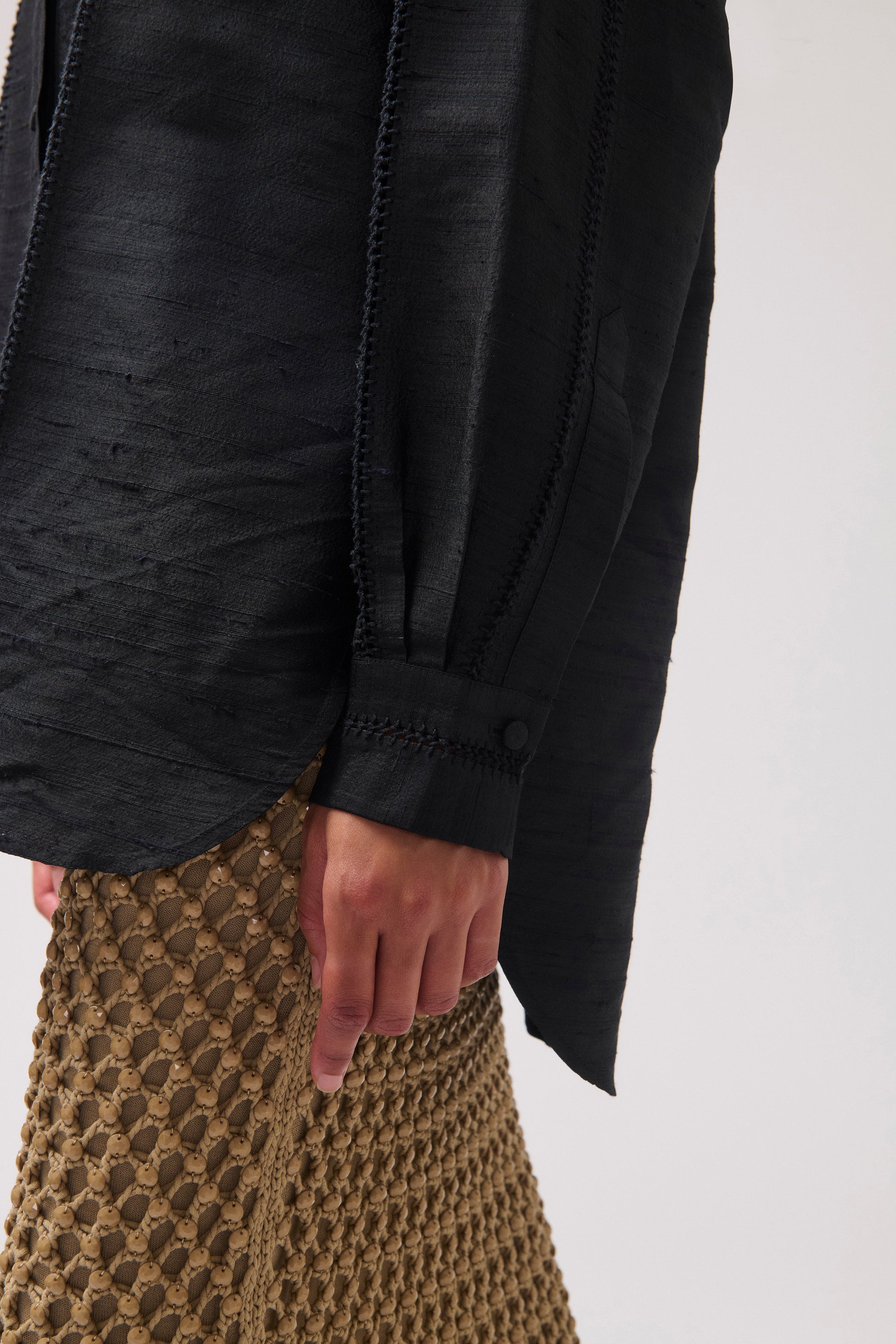 Close-up of a person wearing a black textured long-sleeve blouse with a button cuff and a skirt against a light gray background.