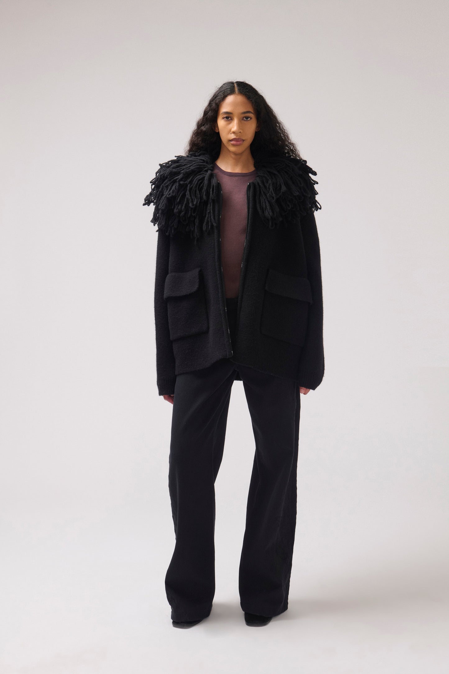 A model stands against a plain off-white studio background wearing a black cropped jacket with an oversized fringe collar and large front patch pockets.
