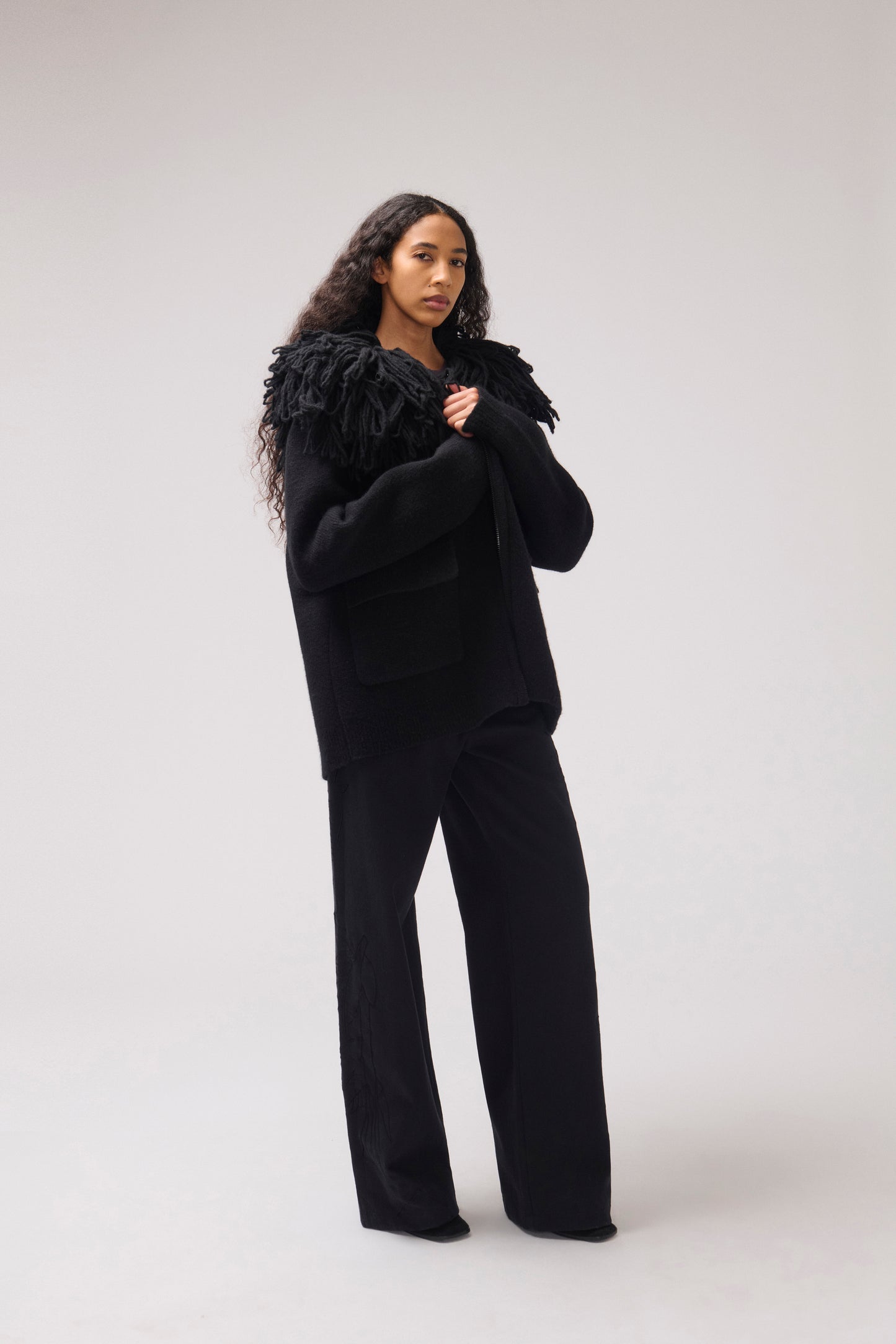 A model stands against a plain off-white studio background wearing a black cropped jacket with an oversized fringe collar and large front patch pockets.