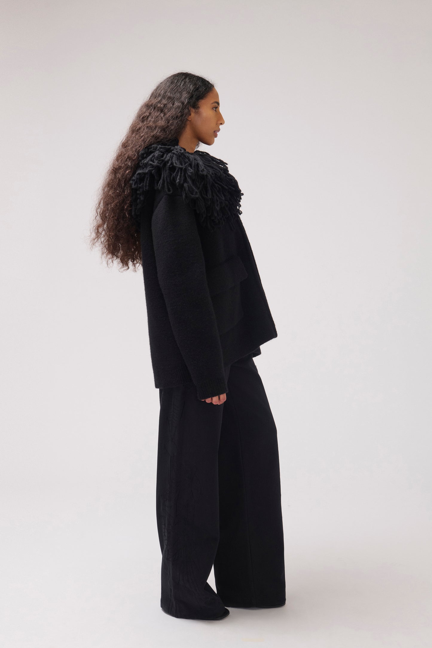 A model standing in profile against a plain off-white studio background wearing a black cropped jacket with an oversized fringe collar and large front patch pockets.