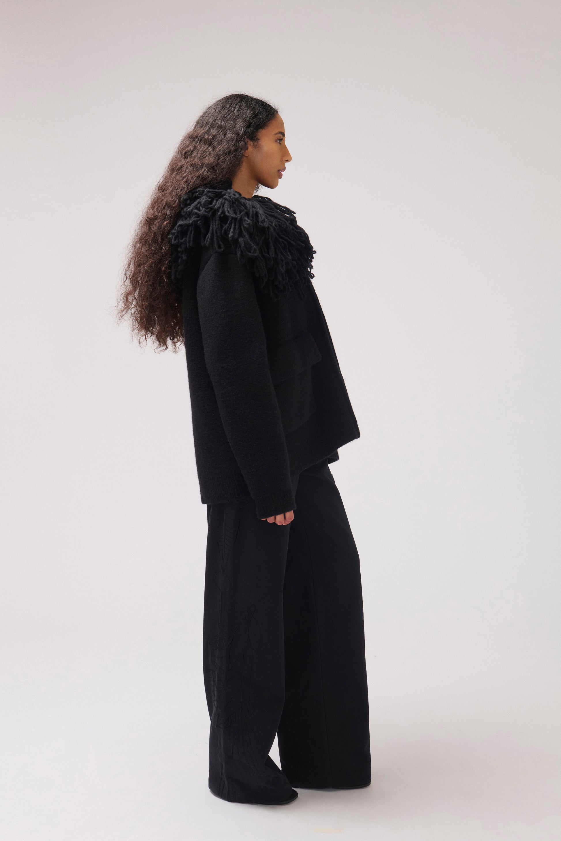 A model standing in profile against a plain off-white studio background wearing a black cropped jacket with an oversized fringe collar and large front patch pockets.