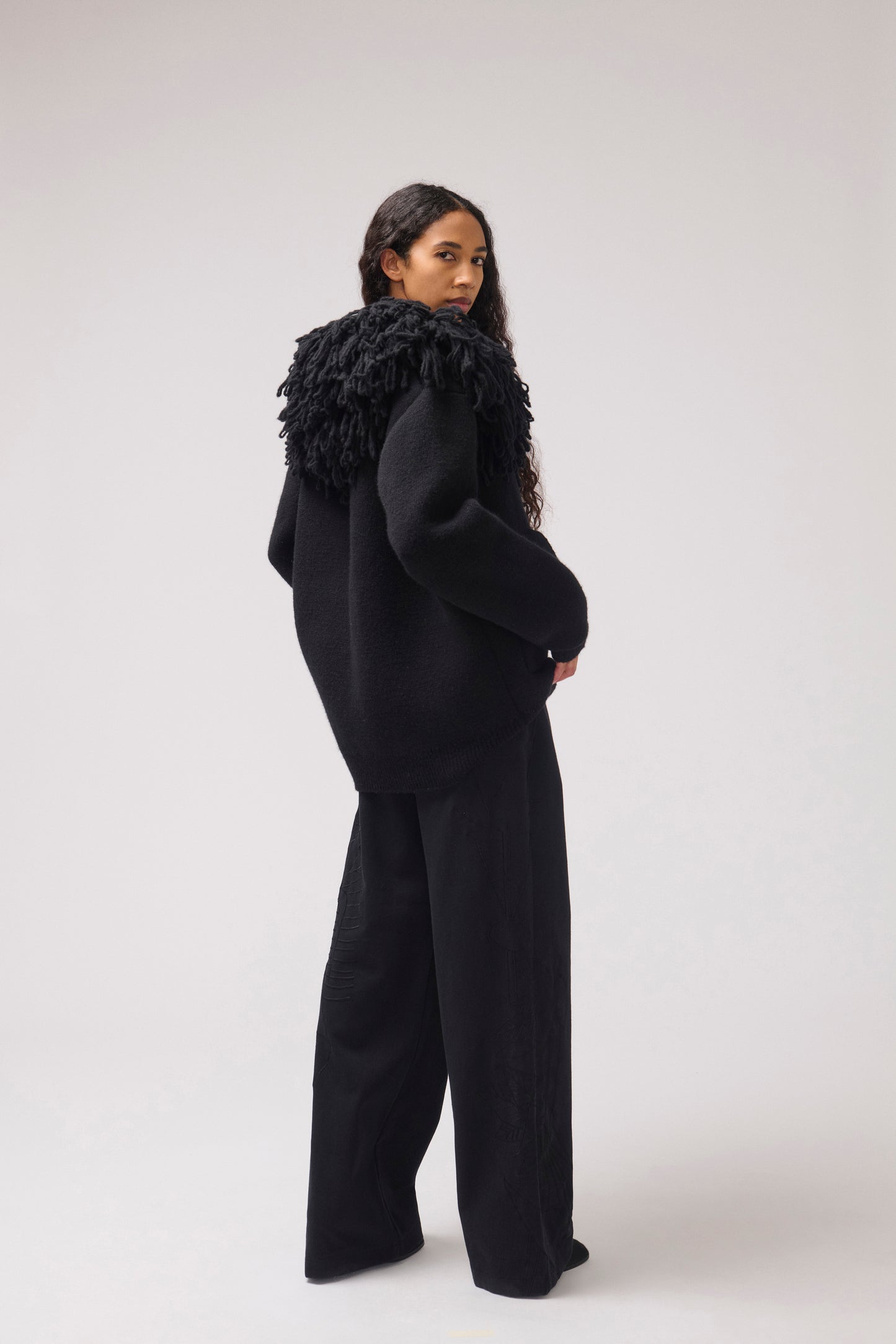 A model looking over her shoulder. She stands against a plain off-white studio background. She is wearing a black cropped jacket with an oversized fringe collar and large front patch pockets. 