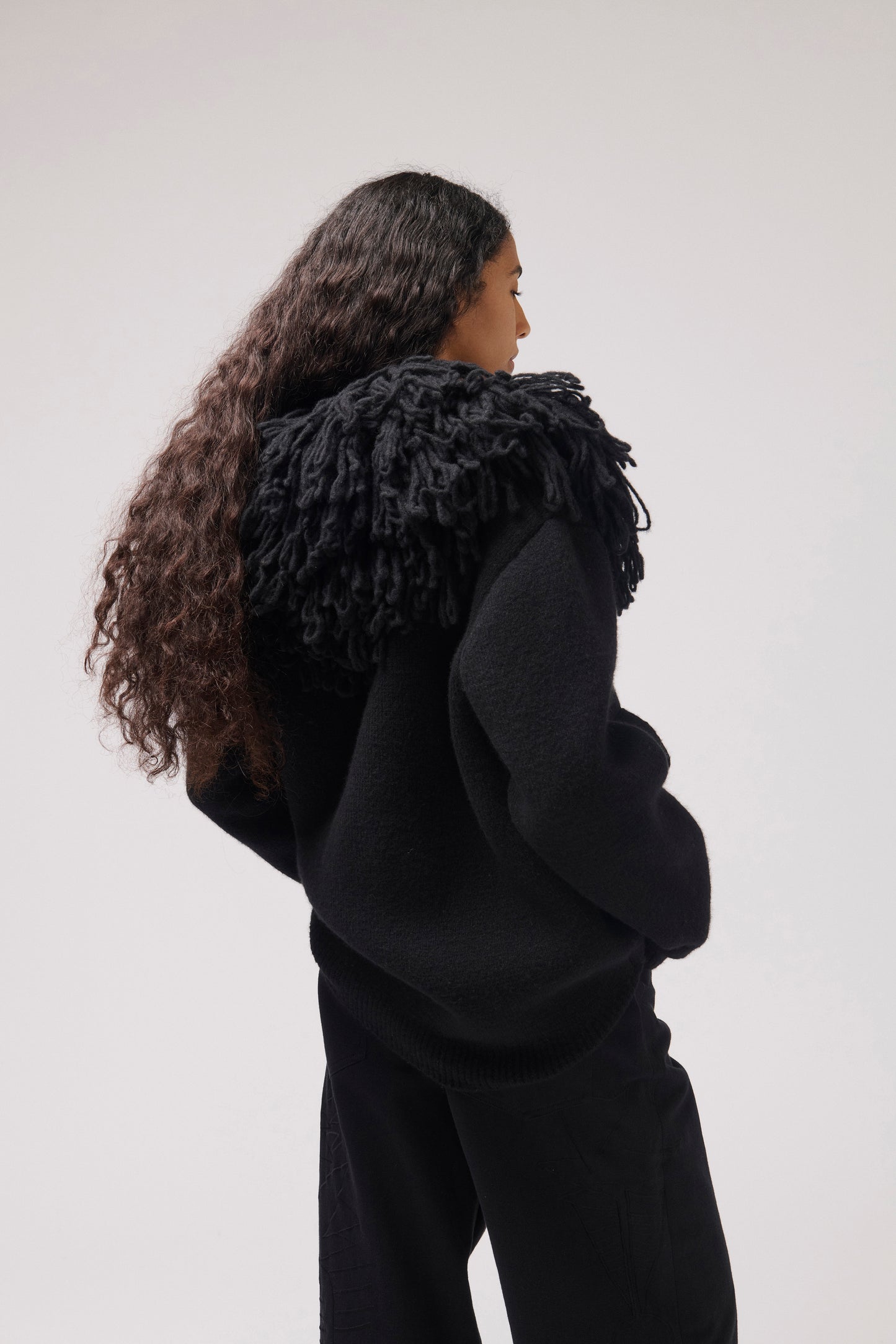 A model shown from the back wearing a black textured knit sweater with a large, shaggy black collar and matching black trousers, set against a plain light gray studio background.