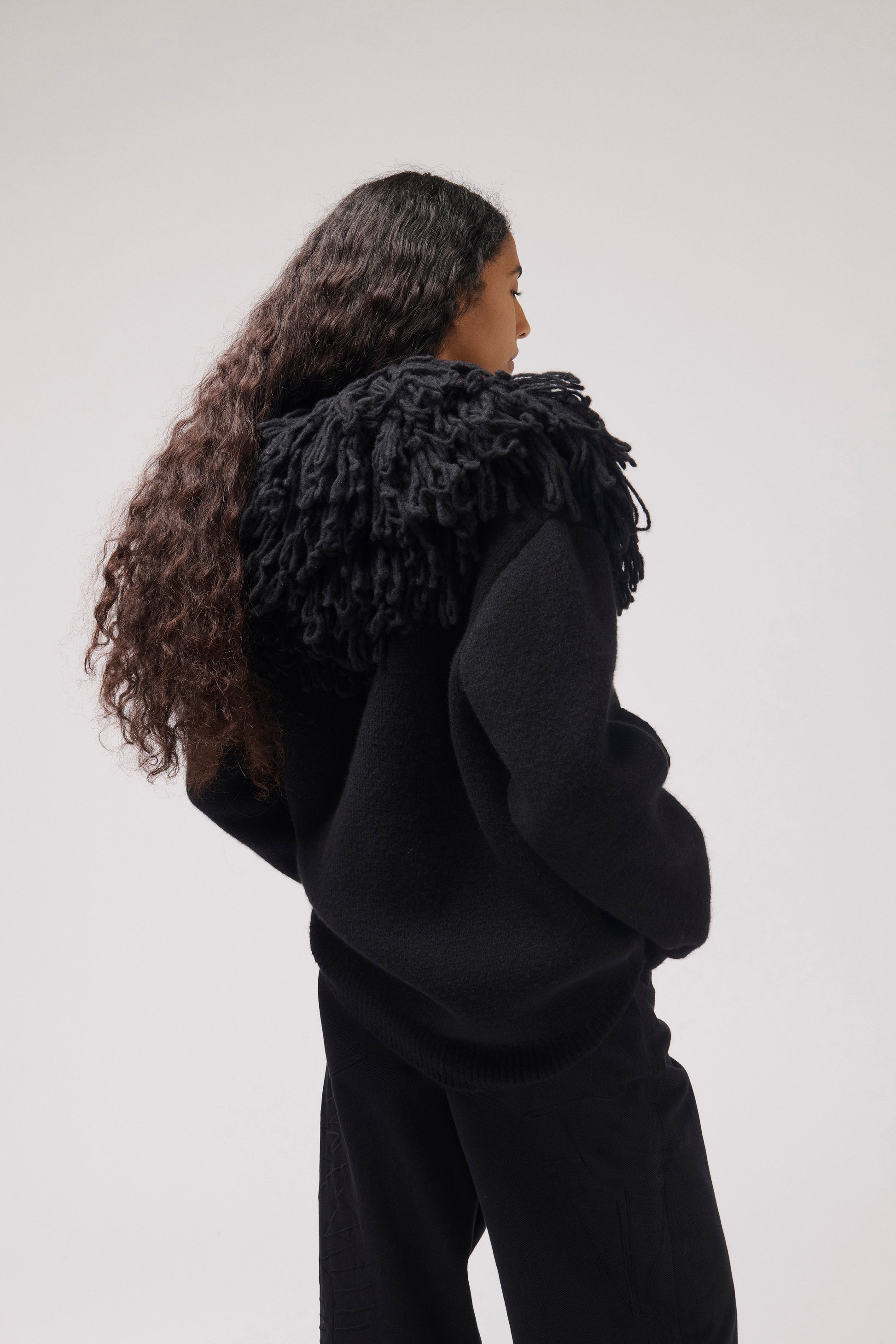 A model shown from the back wearing a black textured knit sweater with a large, shaggy black collar and matching black trousers, set against a plain light gray studio background.