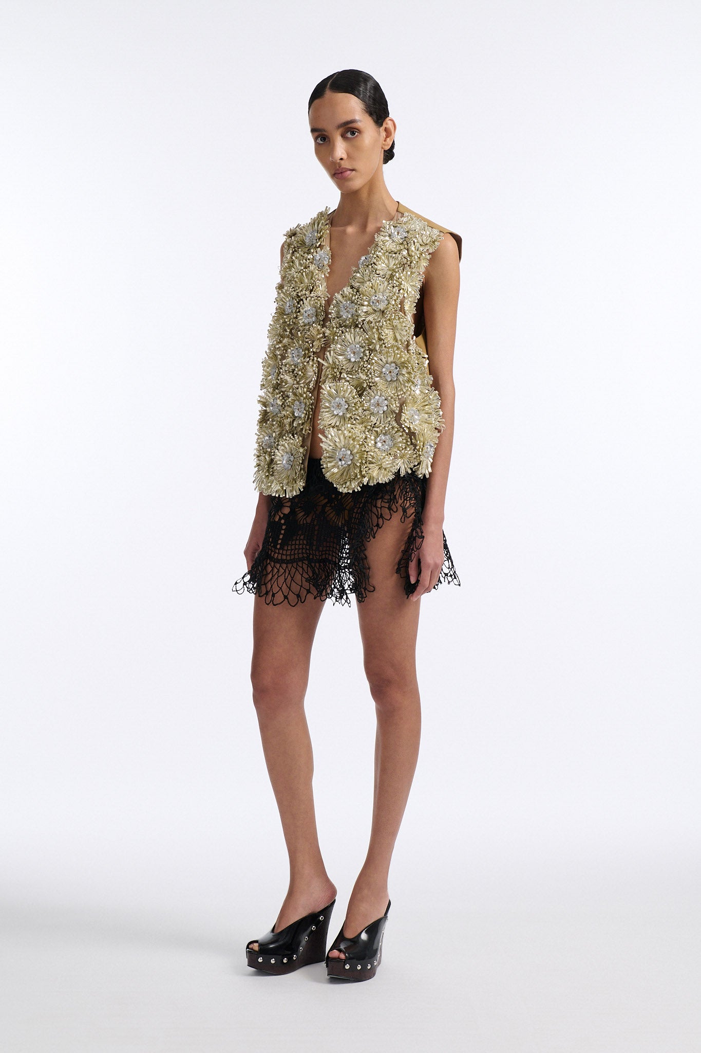 Female model with slicked back black hair in a bun wears metallic ribbon flower V-neck one button vest in pale gold and silver with black see-through crochet mini skirt. 