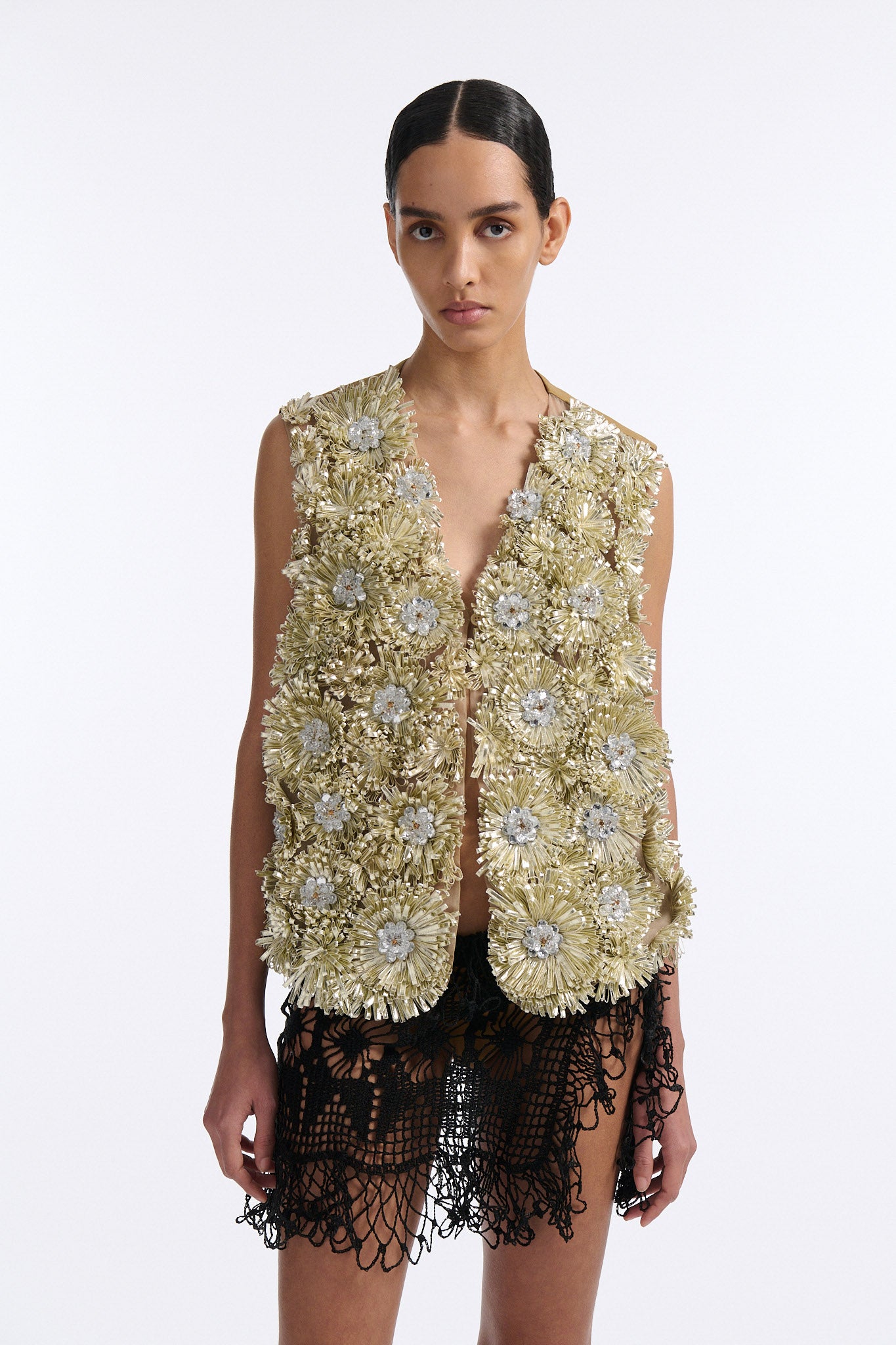 Female model with slicked back black hair in a bun wears metallic ribbon flower V-neck one button vest in pale gold and silver with black see-through crochet mini skirt. 