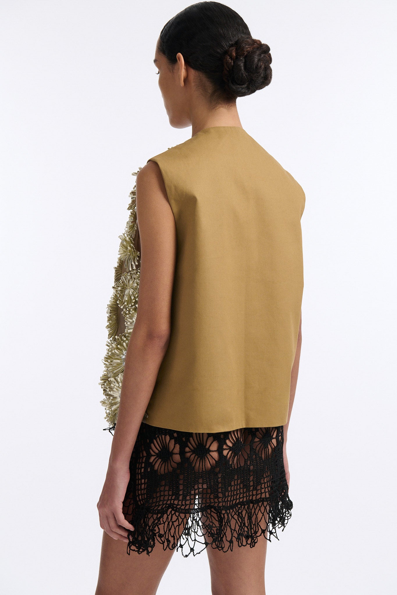 Female model with slicked back black hair in a bun wears metallic ribbon flower V-neck one button vest in pale gold and silver with black see-through crochet mini skirt. 
