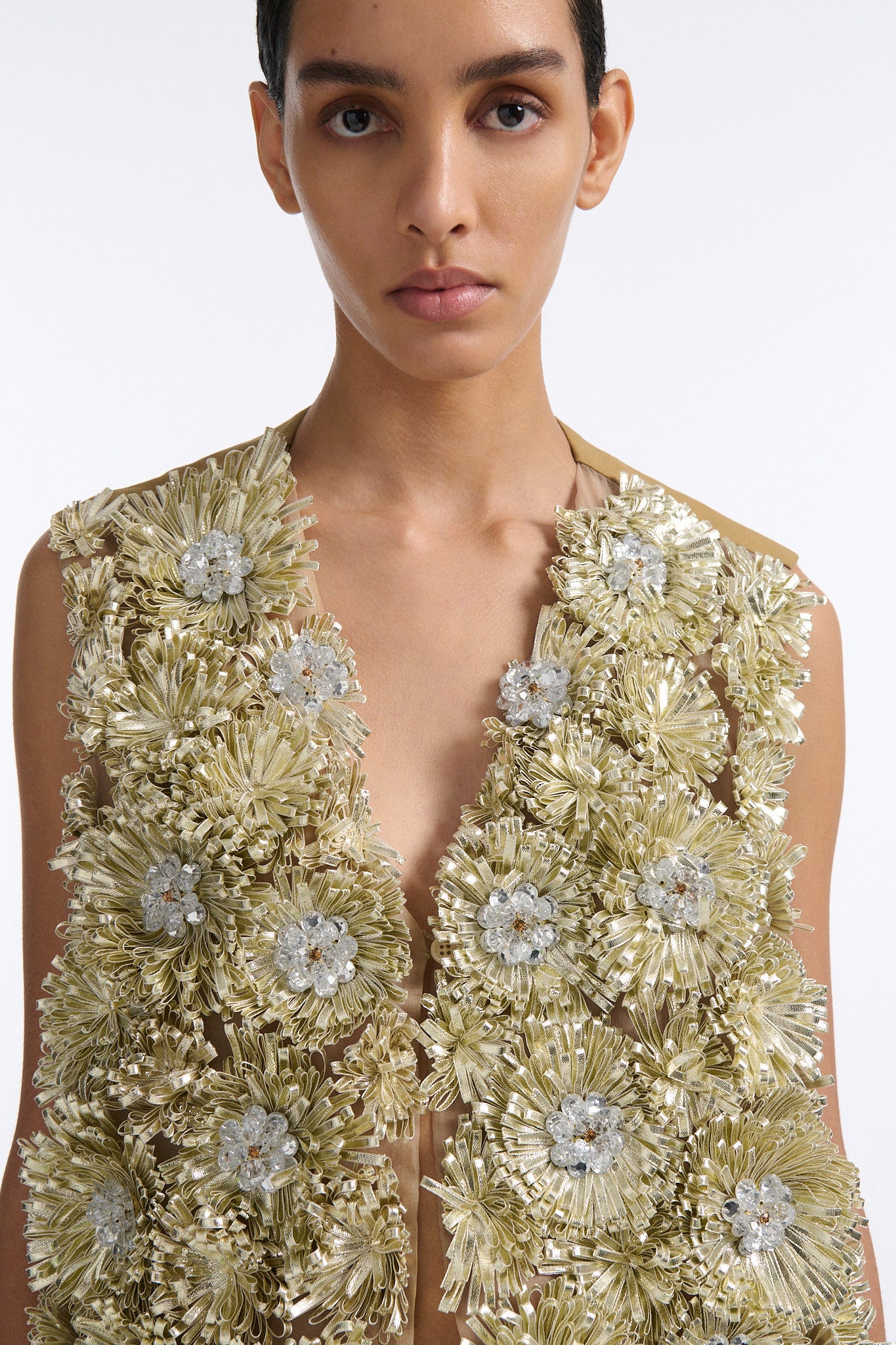 Female model with slicked back black hair in a bun wears metallic ribbon flower V-neck one button vest in pale gold and silver with black see-through crochet mini skirt. 