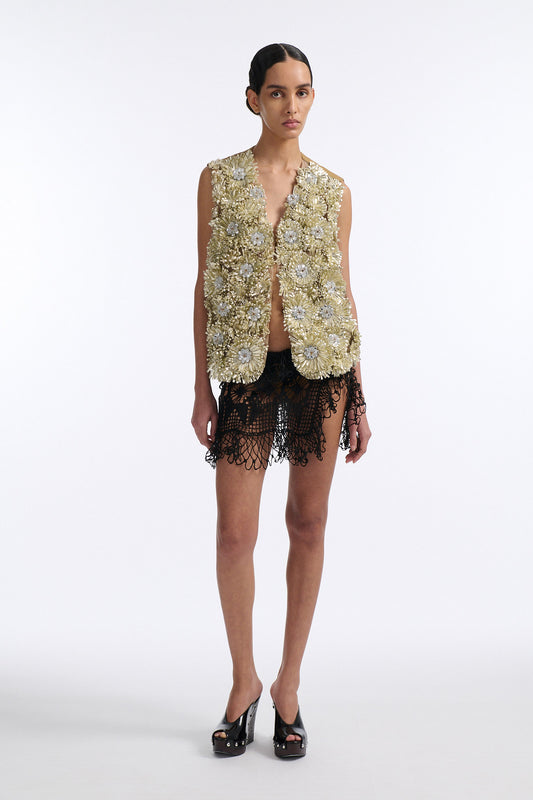 Female model with slicked back black hair in a bun wears metallic ribbon flower V-neck one button vest in pale gold and silver with black see-through crochet mini skirt.