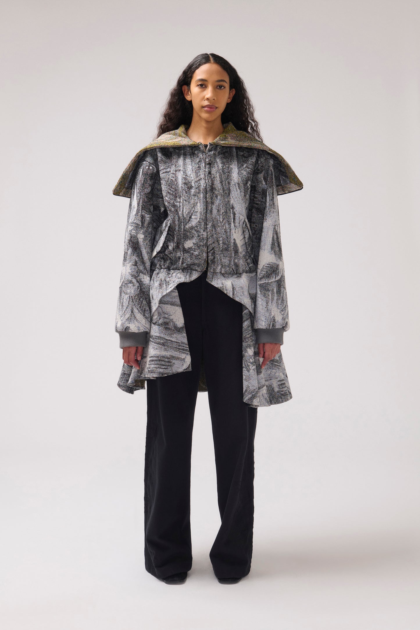 A model stands against a light gray studio background wearing a long-sleeved, asymmetrical jacket with a gray and white abstract print, a wide collar, and black wide-leg pants.