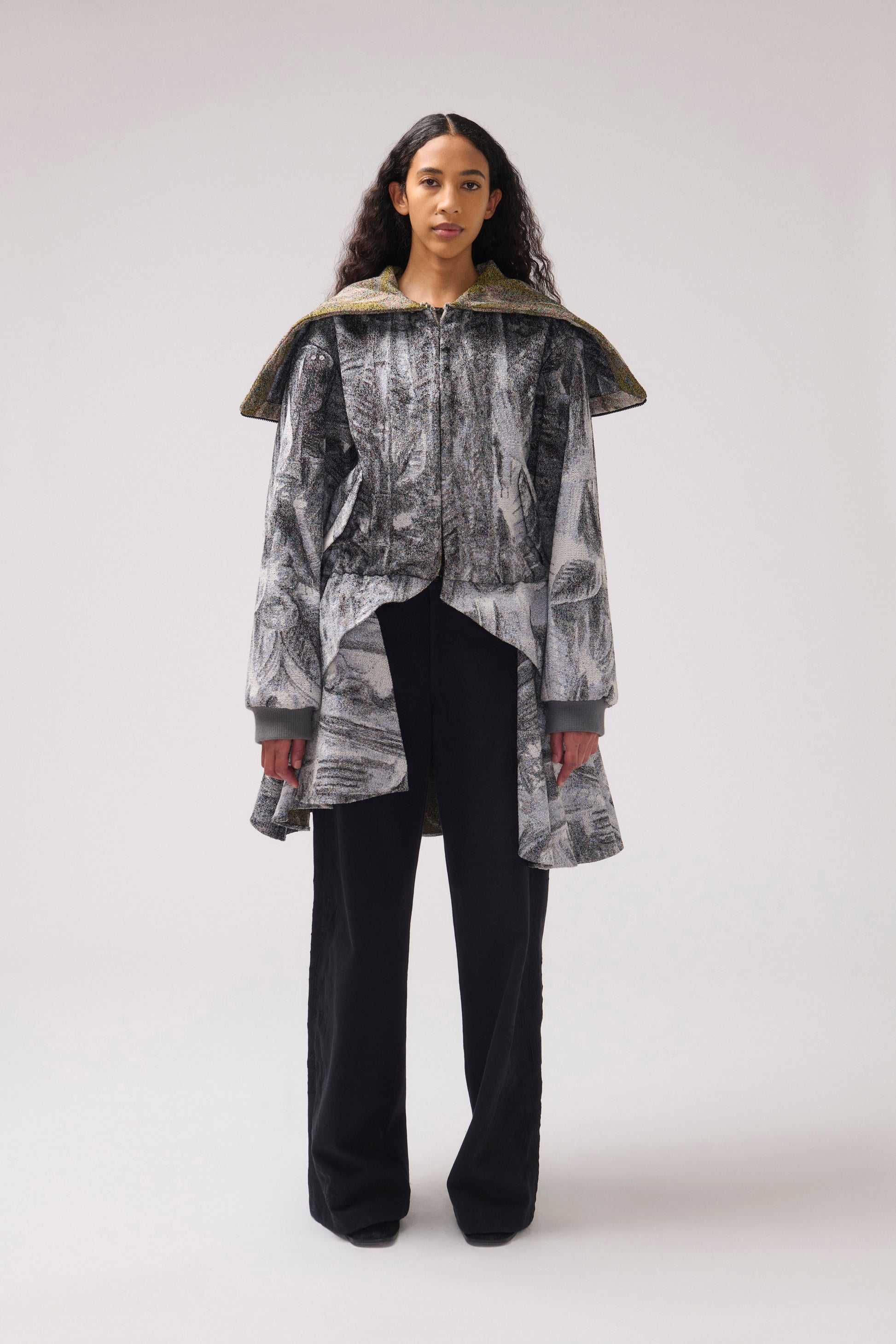 A model stands against a light gray studio background wearing a long-sleeved, asymmetrical jacket with a gray and white abstract print, a wide collar, and black wide-leg pants.