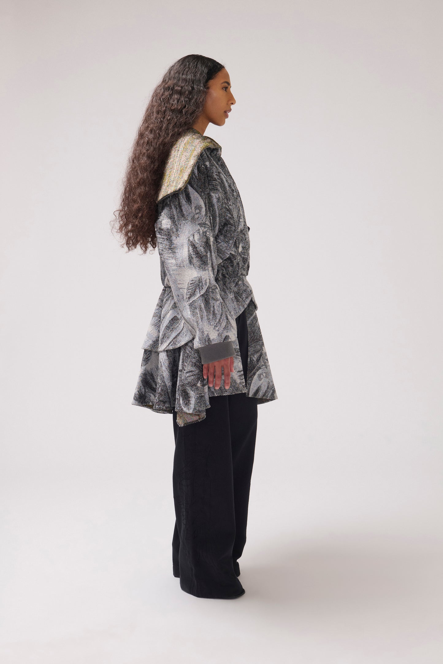 A model pictured in profile wearing a long-sleeved, asymmetrical jacket with a gray and white abstract print, a wide collar ruffle detail at the waist and black wide-leg pants.