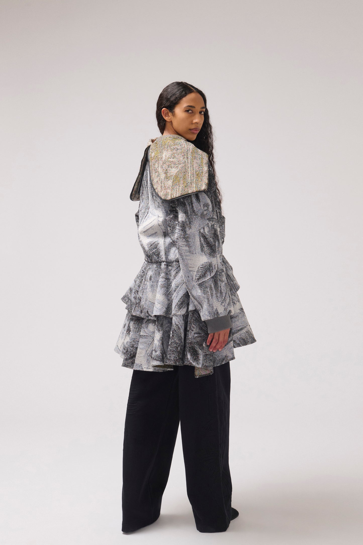 A model is shown looking over her back wearing a tiered, ruffled jacket with a large, wide collar in a gray and black abstract print, paired with black wide-leg trousers, against a plain light gray studio background.