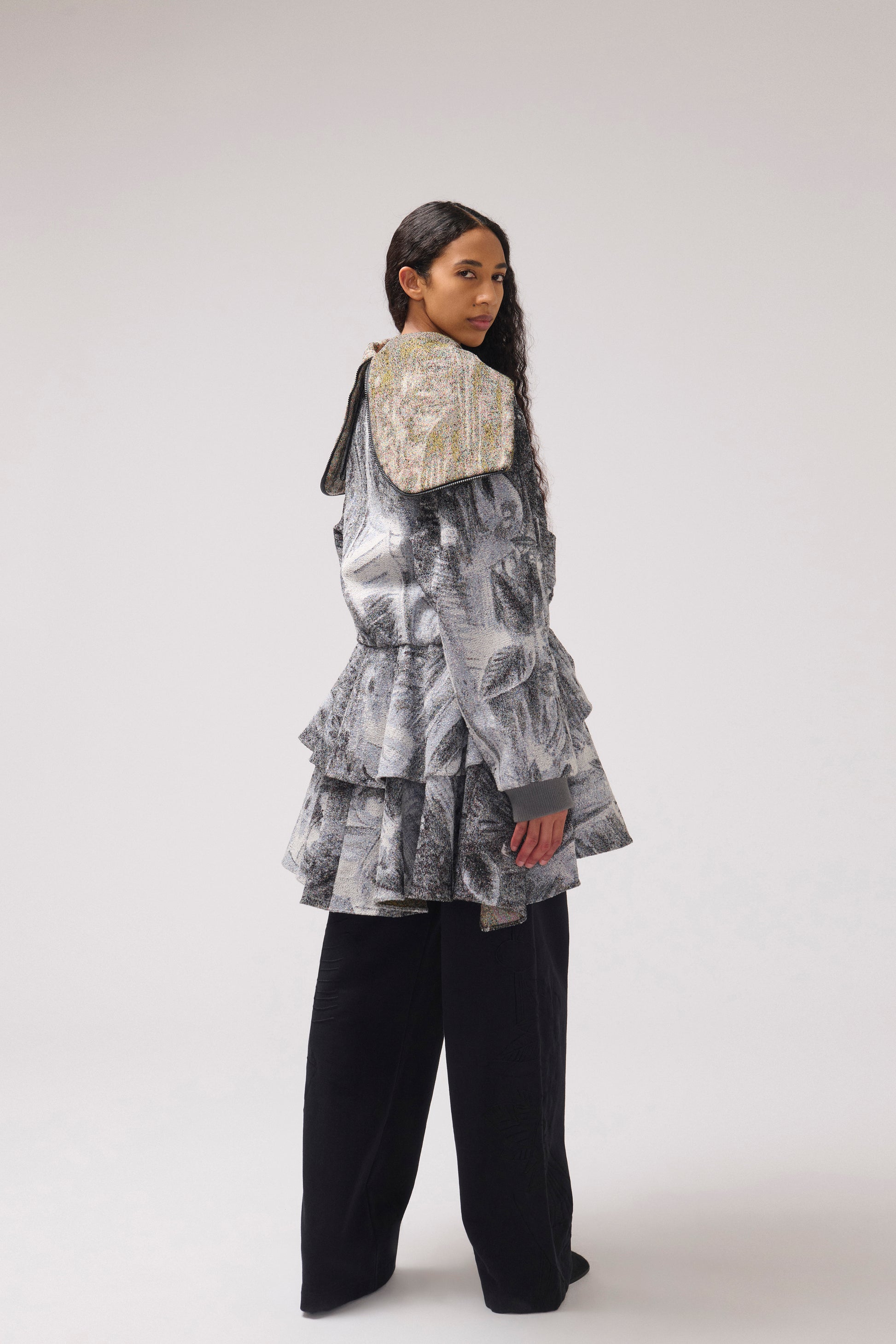 A model is shown looking over her back wearing a tiered, ruffled jacket with a large, wide collar in a gray and black abstract print, paired with black wide-leg trousers, against a plain light gray studio background.