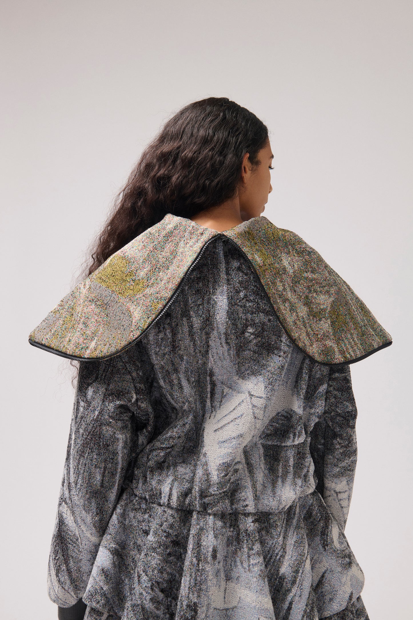 A model shown from the back wearing a long-sleeved, asymmetrical jacket with a gray and white abstract print, and a wide collar.