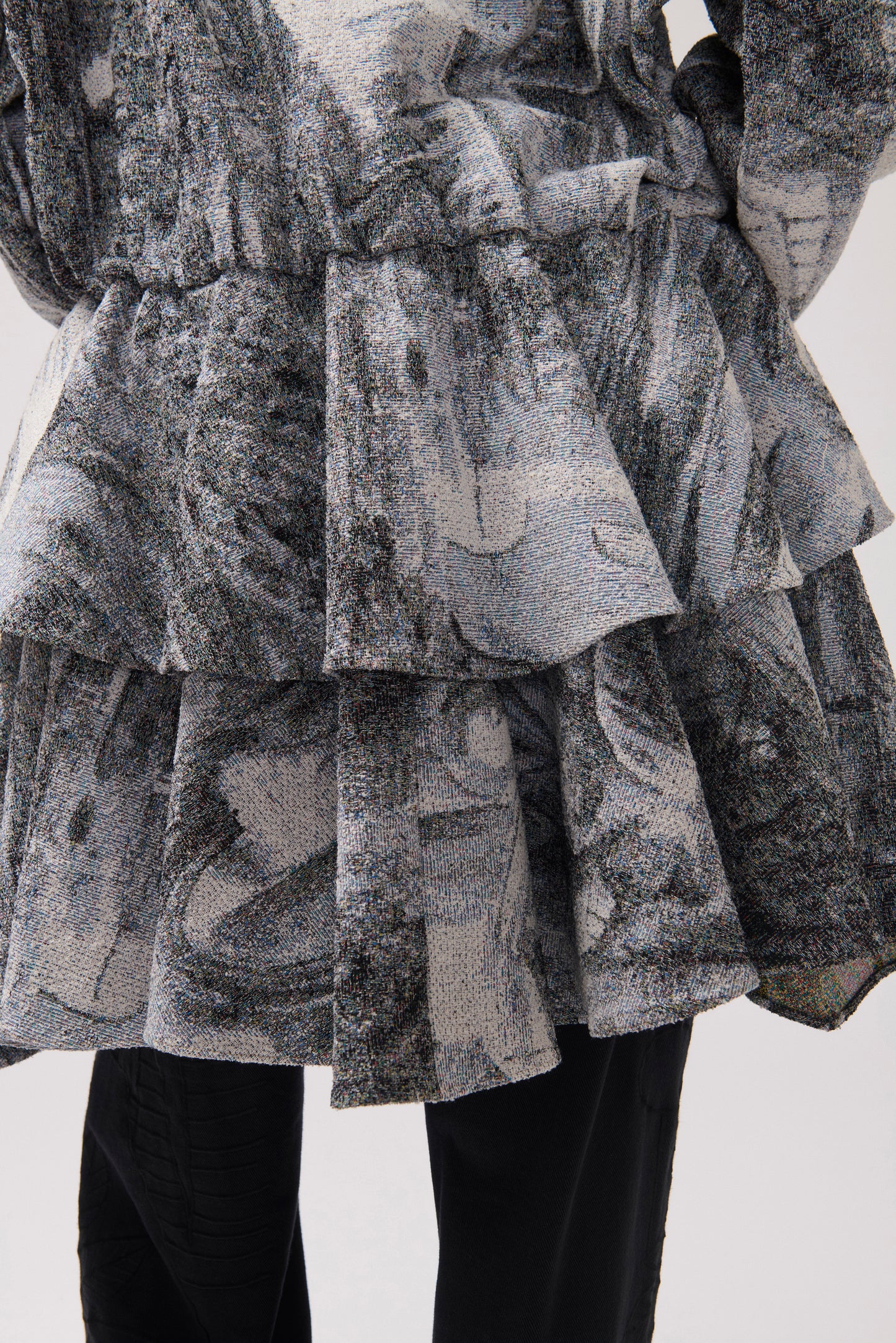 A close-up of a person from behind. The person is wearing a long-sleeved, asymmetrical jacket with a gray and white abstract print, a wide ruffle detail at the waist and black wide-leg pants.