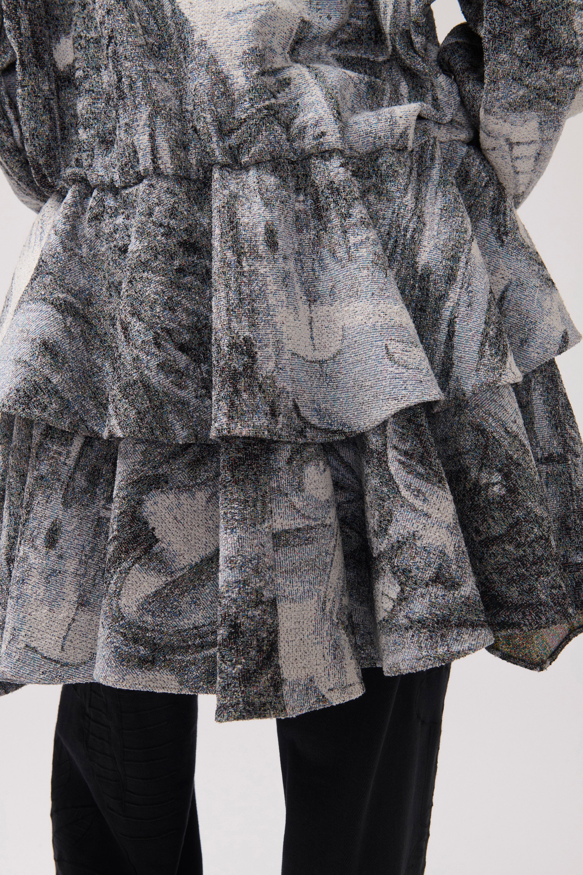 A close-up of a person from behind. The person is wearing a long-sleeved, asymmetrical jacket with a gray and white abstract print, a wide ruffle detail at the waist and black wide-leg pants.