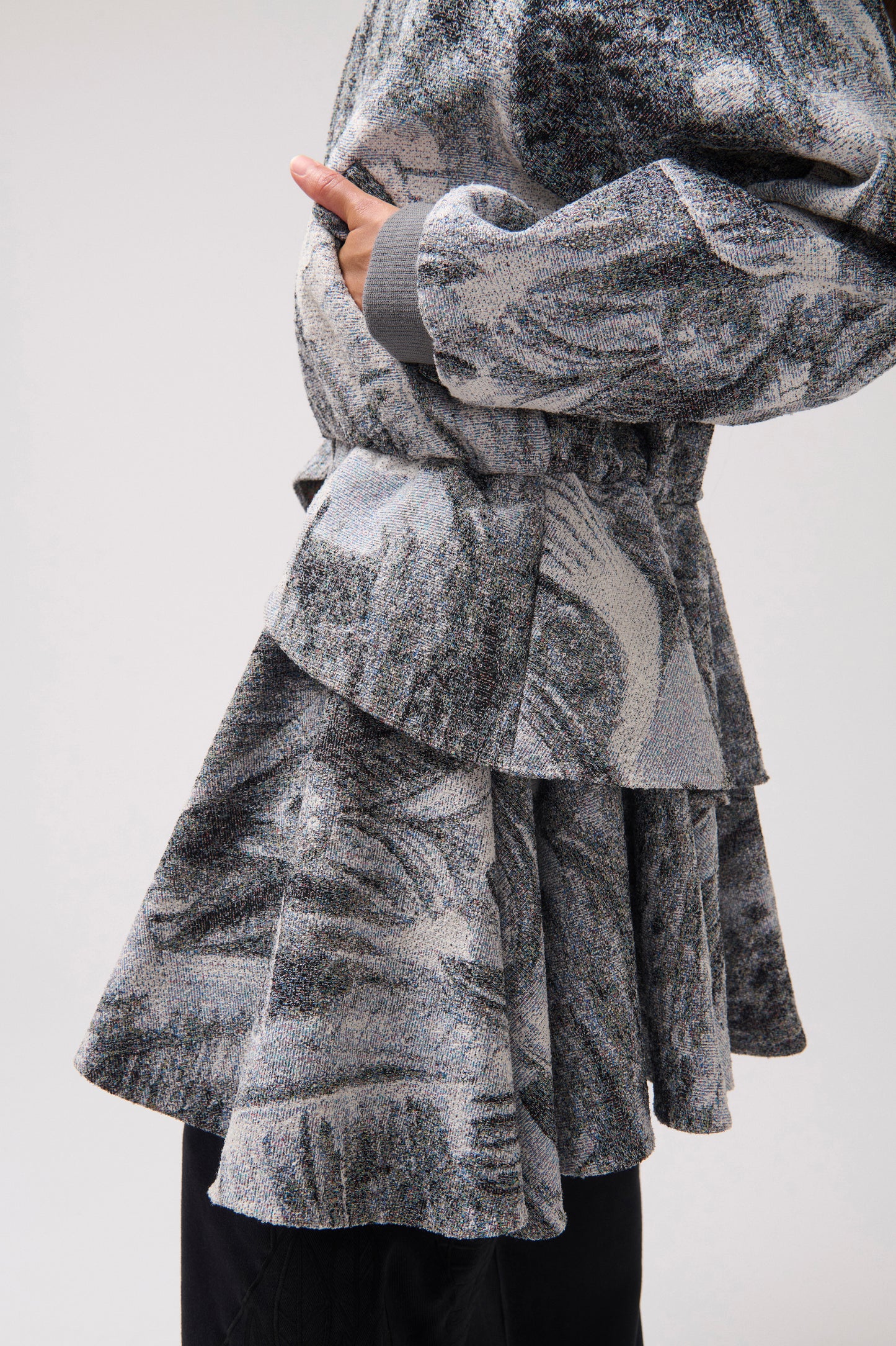 A close-up of a person wearing a long-sleeved, asymmetrical jacket with a gray and white abstract print, a wide ruffle detail at the waist and black wide-leg pants.