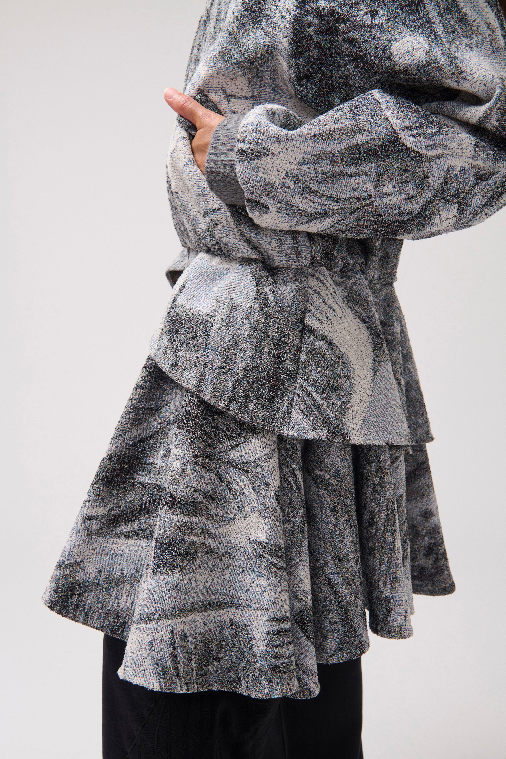 A close-up of a person wearing a long-sleeved, asymmetrical jacket with a gray and white abstract print, a wide ruffle detail at the waist and black wide-leg pants.