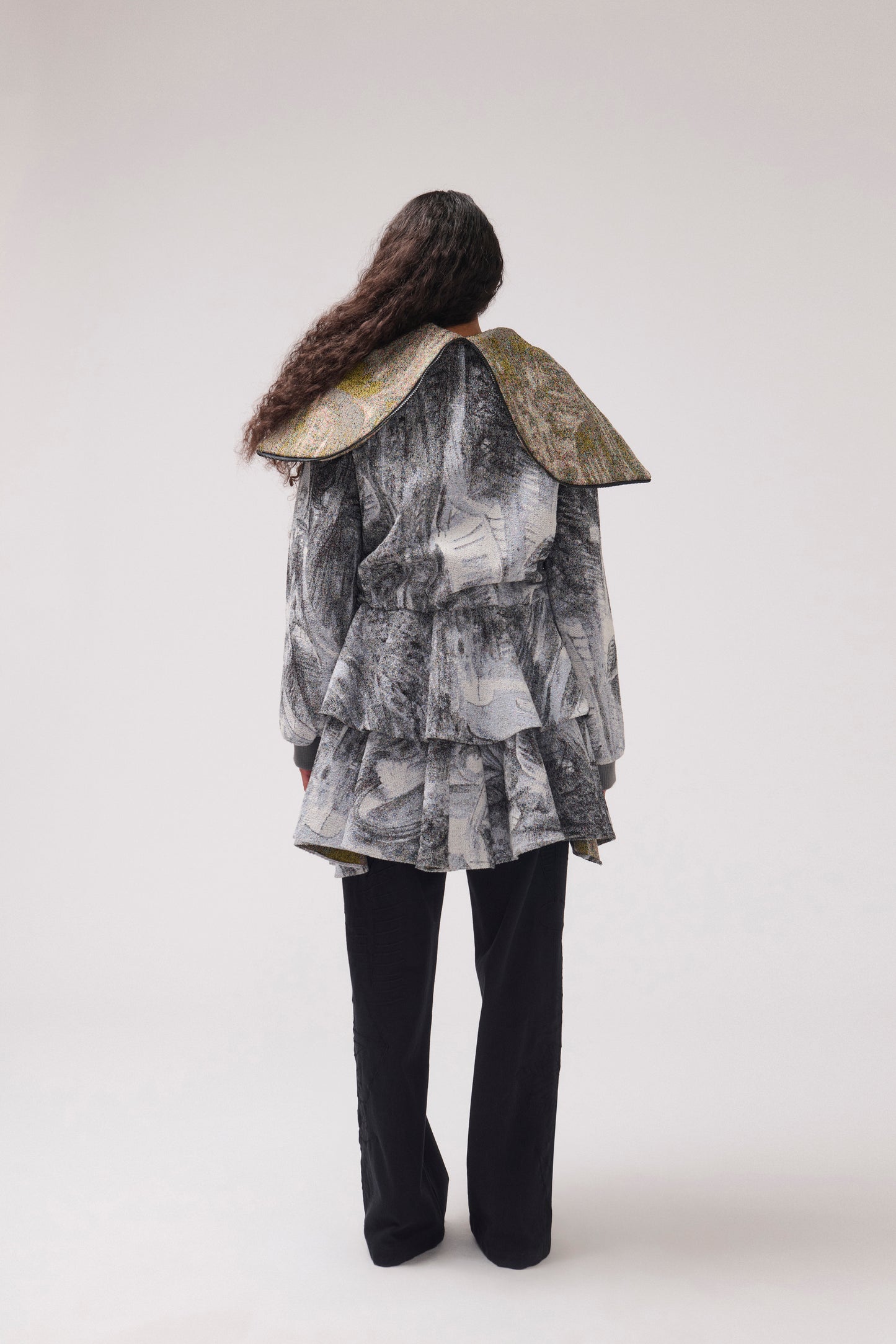 A model viewed from behind wearing a long-sleeved, asymmetrical jacket with a gray and white abstract print, a wide collar ruffle detail at the waist and black wide-leg pants.