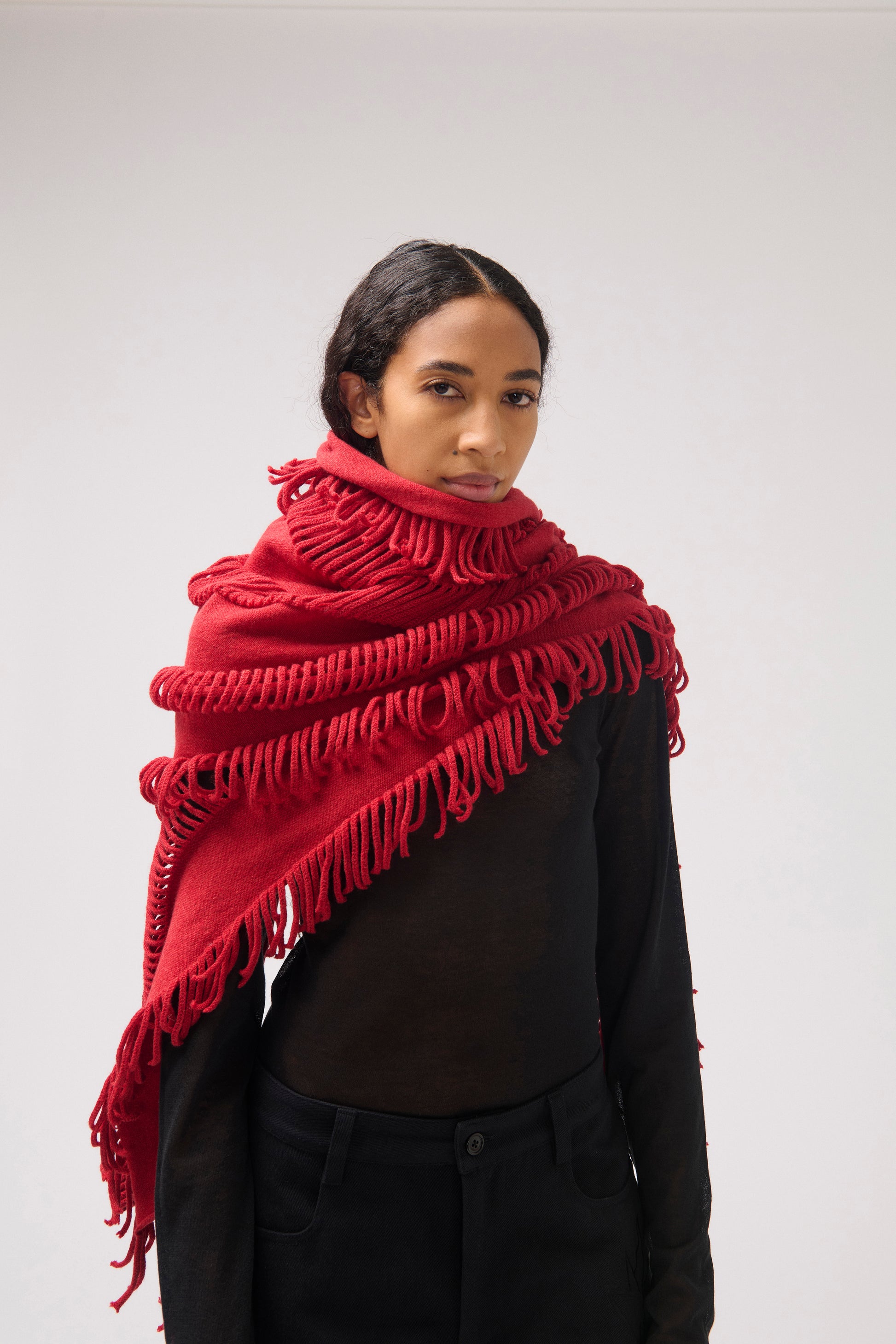 A model wearing a vibrant red fringed shawl over a black top and black pants, photographed against a neutral light gray studio background.
