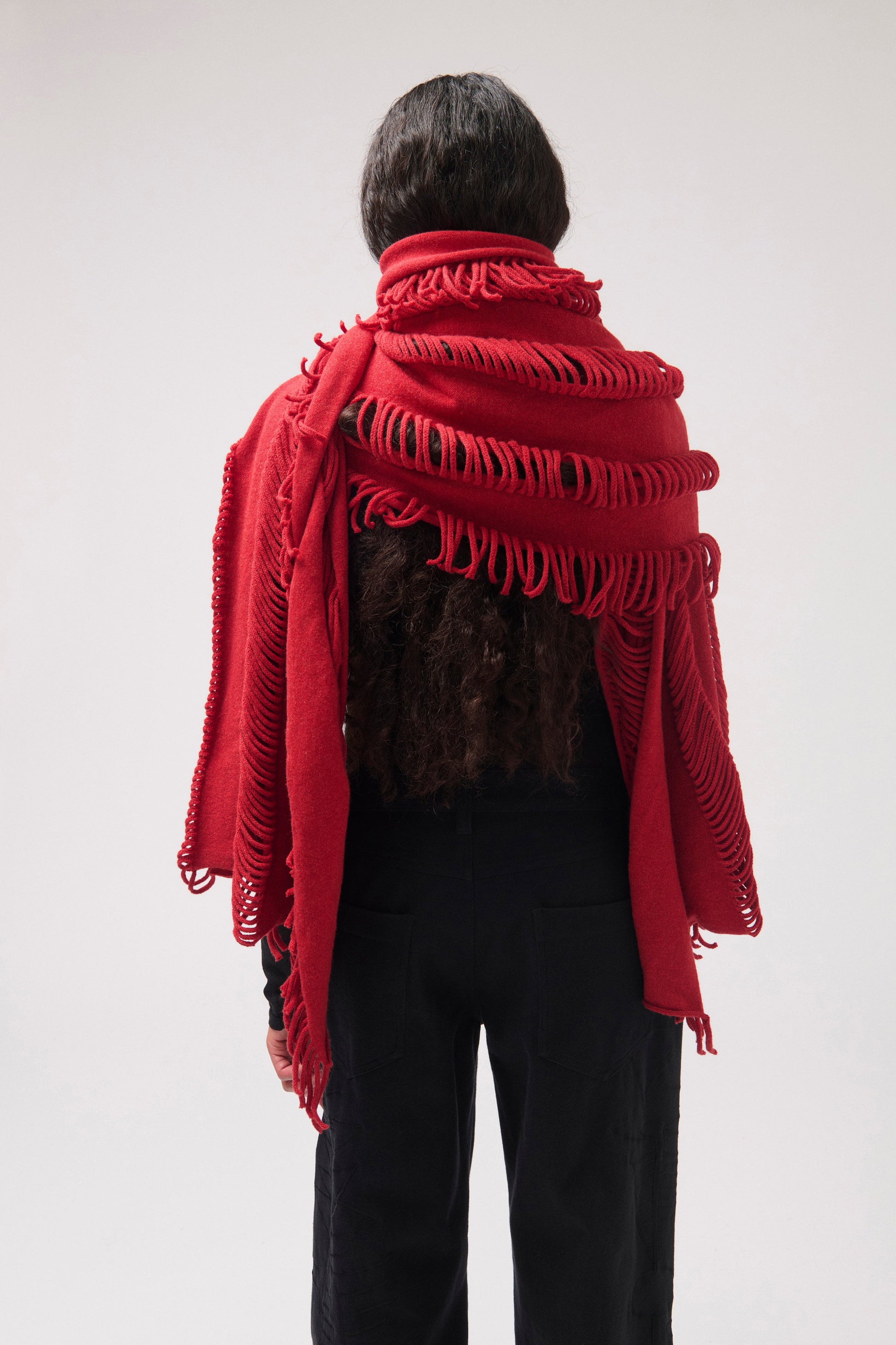 A model photographed from behind wearing a vibrant red fringed shawl over a black top and black pants, photographed against a neutral light gray studio background.