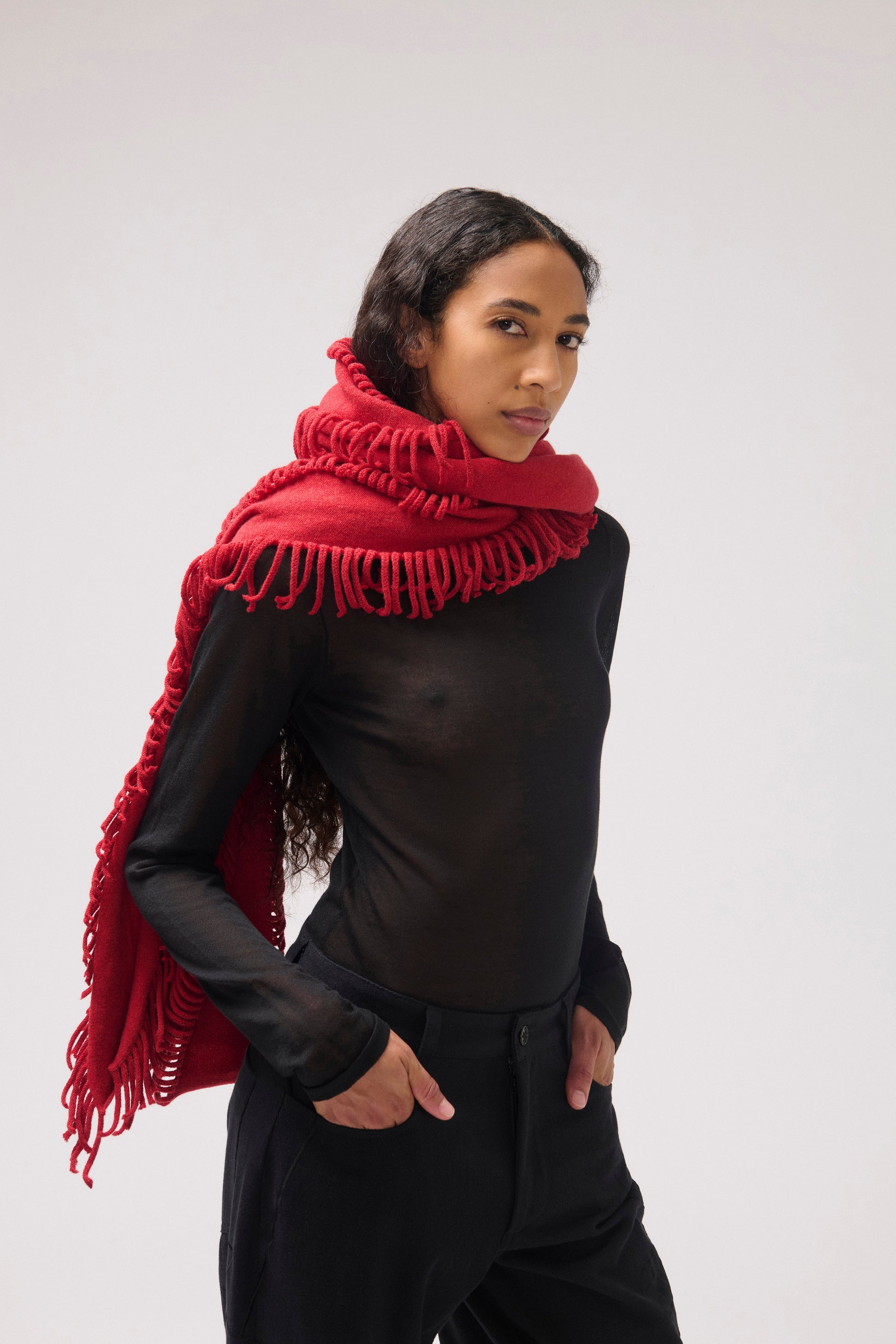 A model wearing a vibrant red fringed shawl over a black top and black pants, photographed against a neutral light gray studio background.